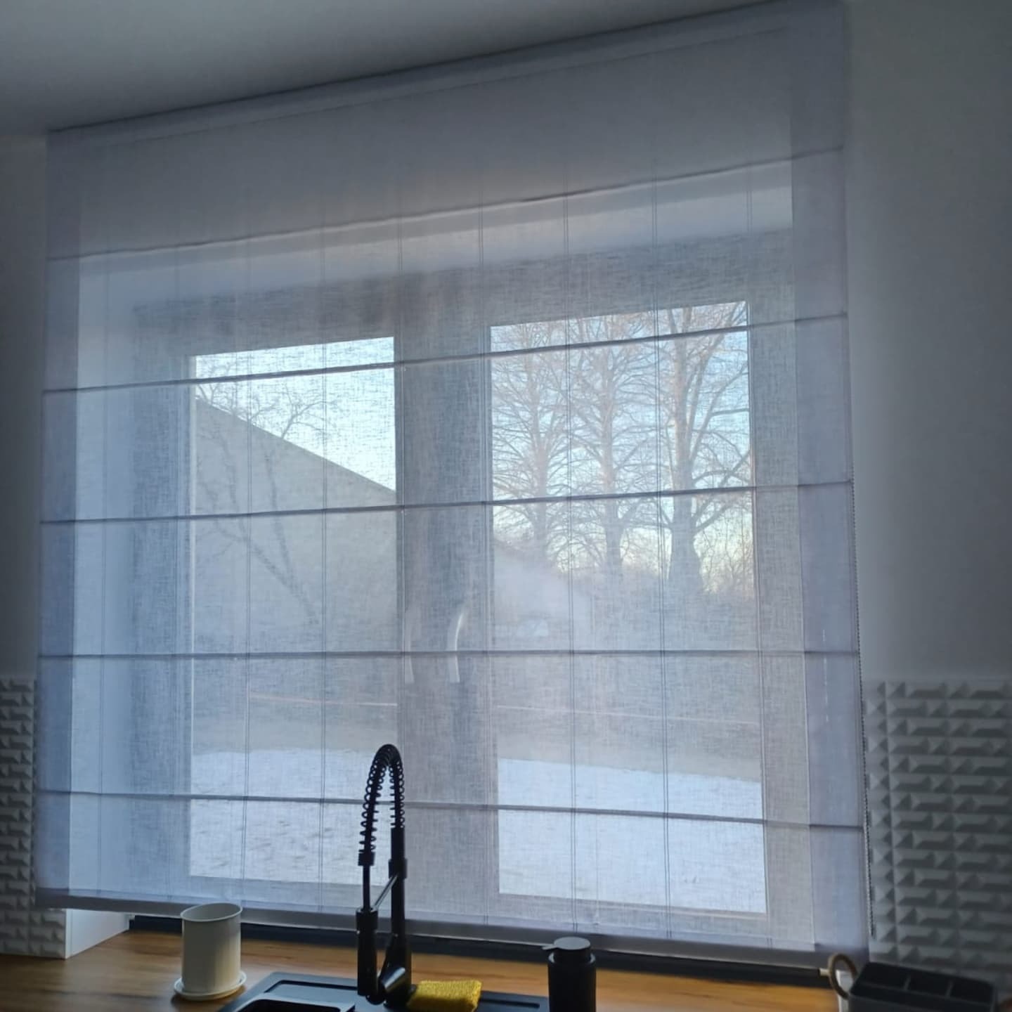 Delikatna, półprzezroczysta roleta w stylu rzymskim zamontowana w oknie kuchennym, widoczna armatura zlewozmywakowa i fragment ściany z dekoracyjną fakturą.