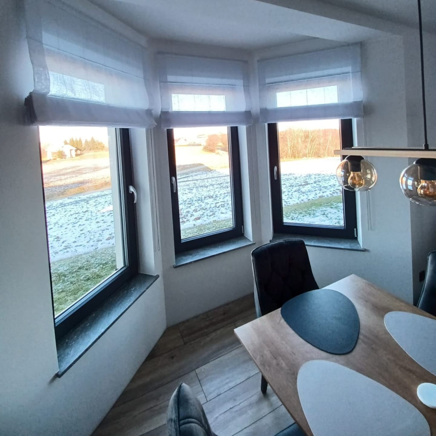 Jasne rolety rzymskie zamontowane w trzech oknach z widokiem na zaśnieżone pole, minimalistyczny wystrój wnętrza z drewnianym stołem i krzesłami.