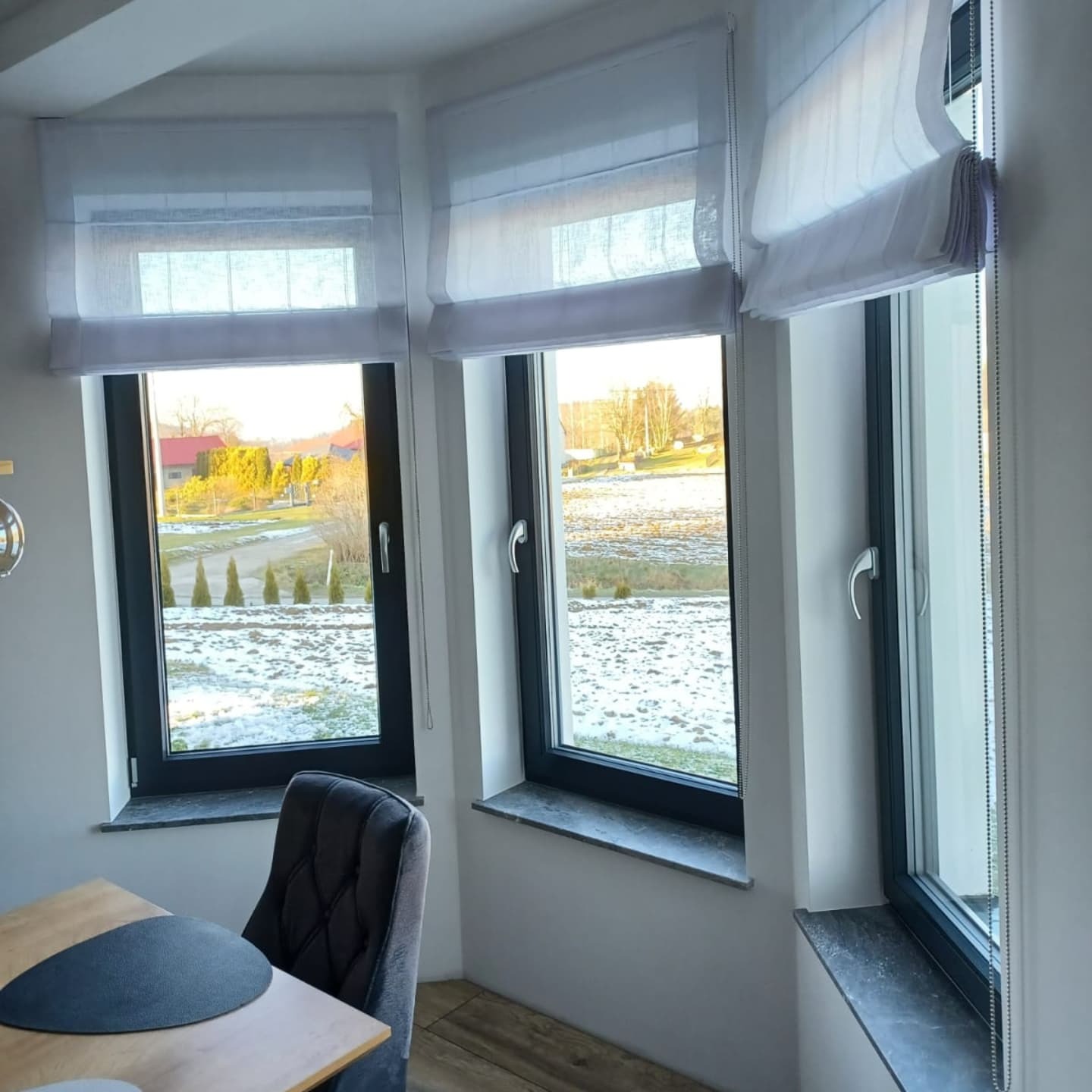 Trzy okna z zamontowanymi białymi roletami rzymskimi, widok na zaśnieżony krajobraz, w tle budynki i drzewa.