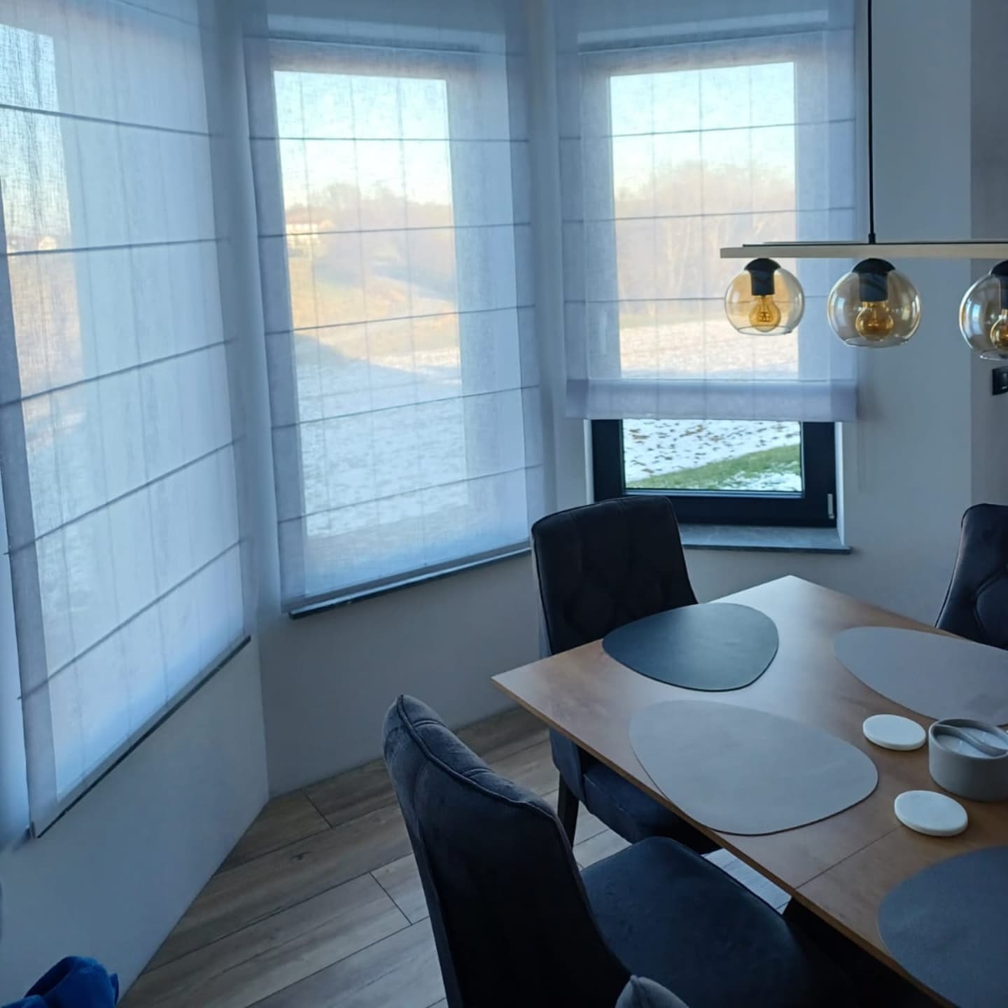 Jasne rolety rzymskie zamontowane w oknach narożnych w jadalni z drewnianym stołem i szarymi krzesłami, widok na zewnątrz z zaśnieżonym krajobrazem.