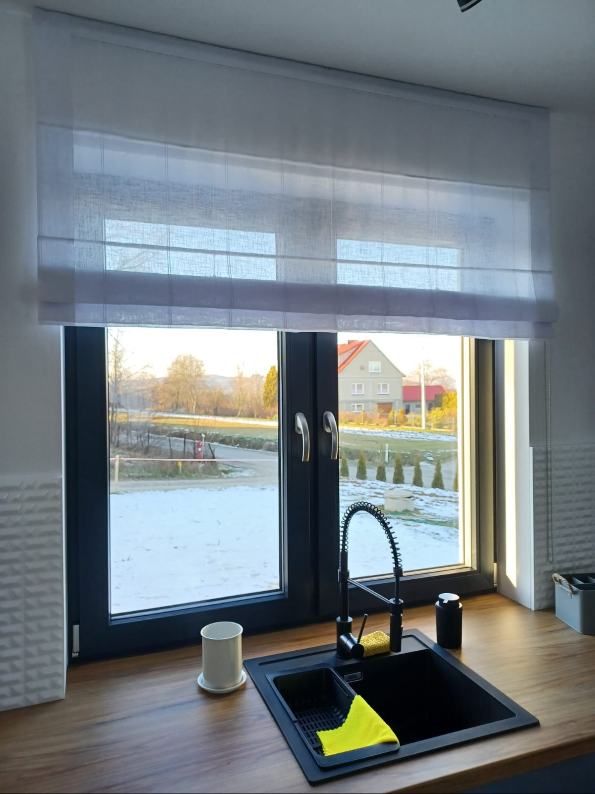 Jasna, półprzezroczysta roleta rzymska w oknie kuchennym z widokiem na zimowy krajobraz za oknem, czarny zlew i bateria kuchenna na pierwszym planie.