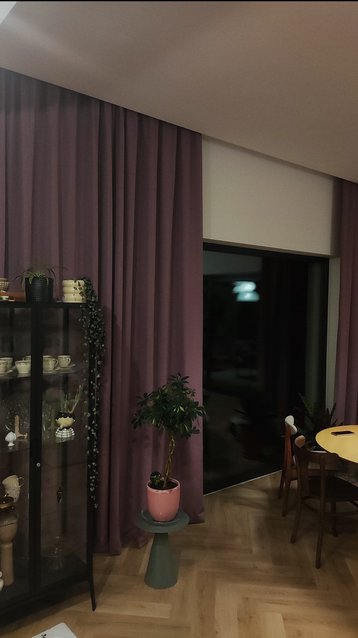 Fioletowe zasłony w salonie z parkietem w jodełkę, obok przeszklona witryna z dekoracjami i rośliny doniczkowe. Widok na okno.