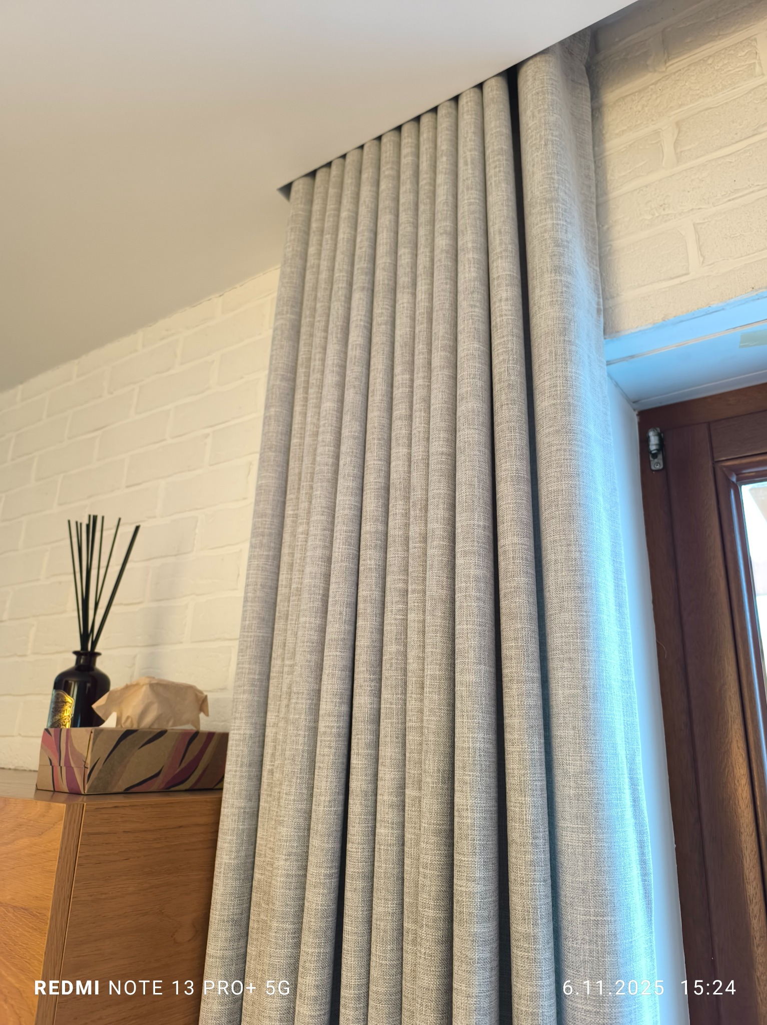 Elegancka, szara zasłona z delikatną fakturą, upięta przy oknie z drewnianą ramą. Na komodzie obok dekoracje: patyczki zapachowe i pudełko.