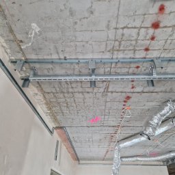 SERPENS COMPANY PROSTA SPÓŁKA AKCYJNA - Betonowy sufit w trakcie montażu instalacji wentylacyjnej z widocznymi metalowymi profilami, kanałami wentylacyjnymi w srebrnej izolacji oraz czerwonymi oznaczeniami na suficie.