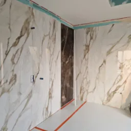 Wnętrze łazienki w trakcie wykańczania: ściany wyłożone białymi płytkami imitującymi marmur z brązowymi żyłami, jedna ściana wyłożona ciemnobrązowymi płytkami, widoczne wystające rury i puszki...