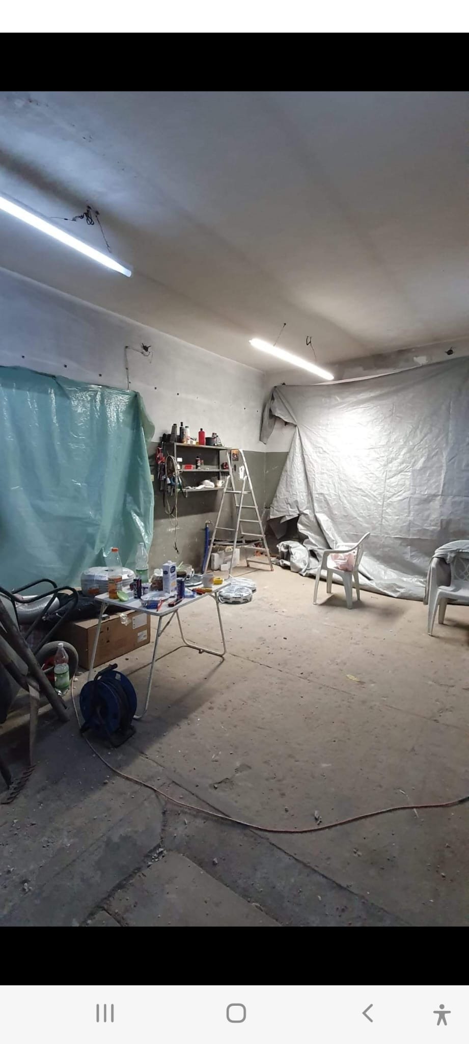 Przygotowania do remontu: pomieszczenie z podłogą betonową, zabezpieczone folią ściany, rozstawiona drabina, stolik z narzędziami i materiałami budowlanymi, widoczne kable i wózek budowlany.