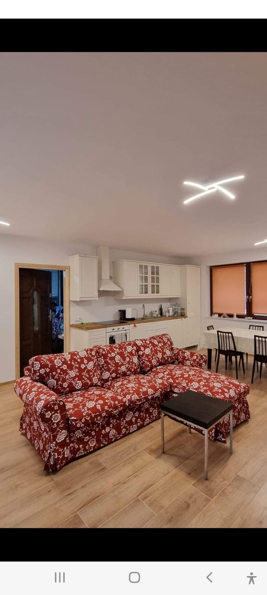 Jasne wnętrze pokoju z drewnopodobną podłogą, sofą w czerwone kwiaty, aneksem kuchennym z białymi szafkami i stołem z krzesłami pod oknem.