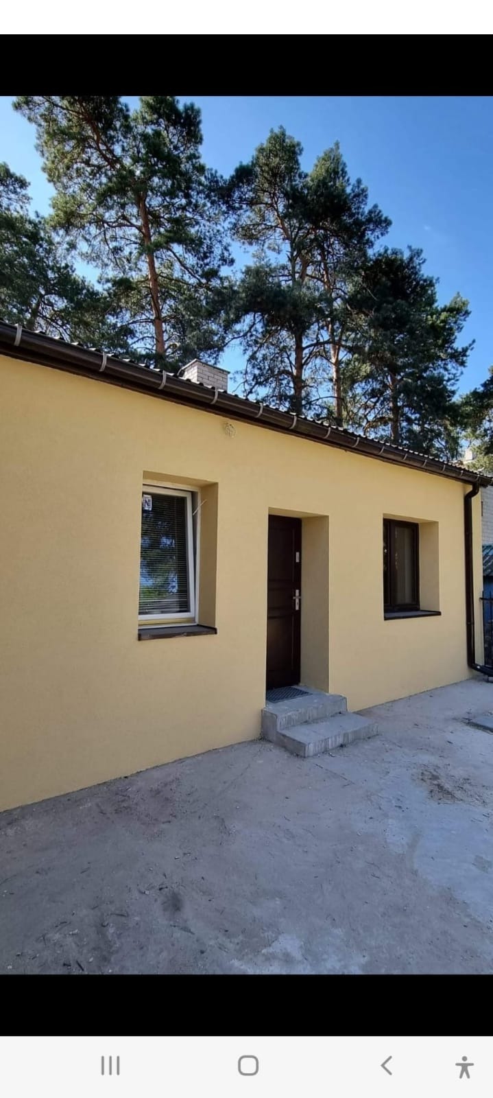 Elewacja budynku jednorodzinnego w odcieniach beżu, z brązowymi elementami wykończeniowymi, widoczne okna z roletami i ciemne drzwi wejściowe, na tle drzew i błękitnego nieba.