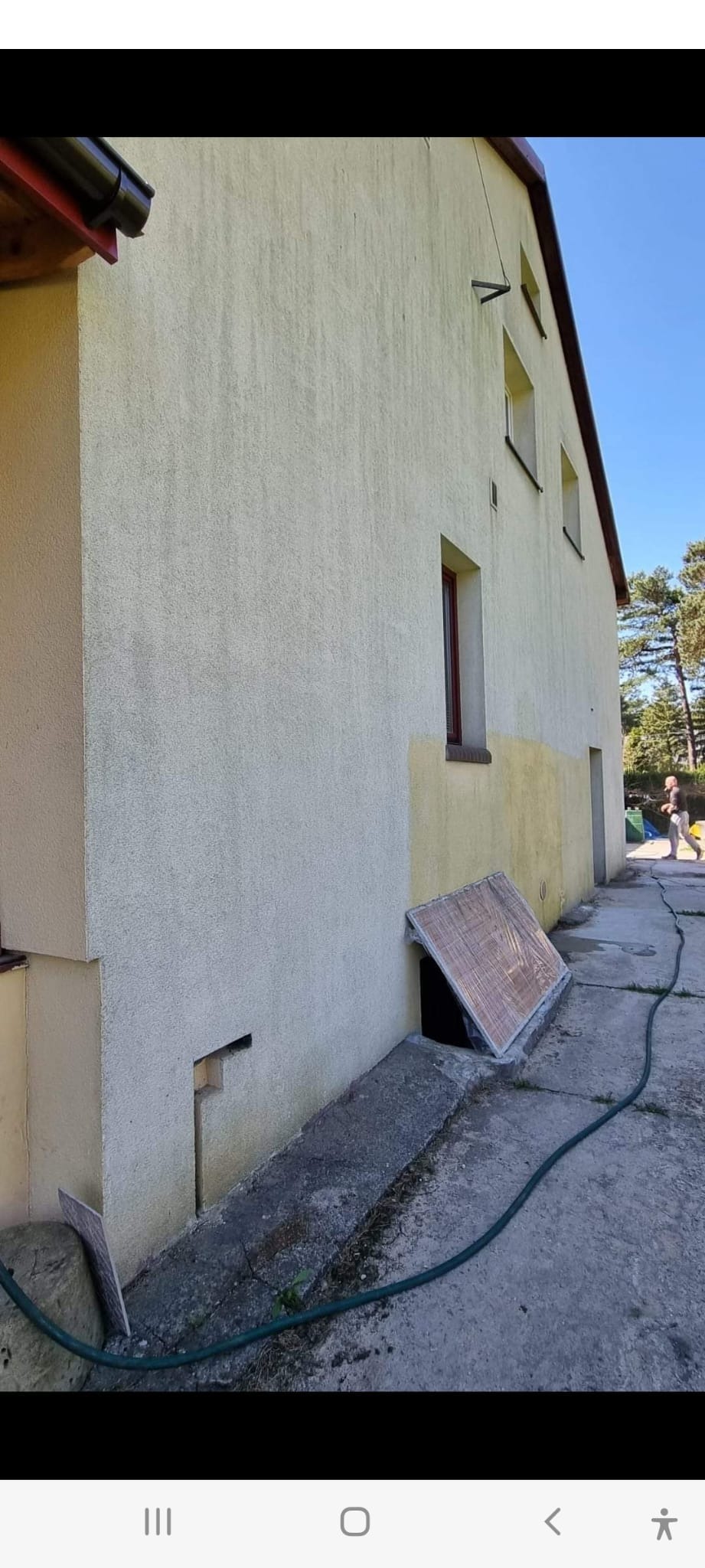 Elewacja dwukondygnacyjnego budynku z fragmentami świeżo nałożonego tynku w odcieniu beżu, widoczne okna z brązowymi ramami, na dole zabezpieczone okno piwniczne, na zewnątrz betonowy chodnik i wąż...