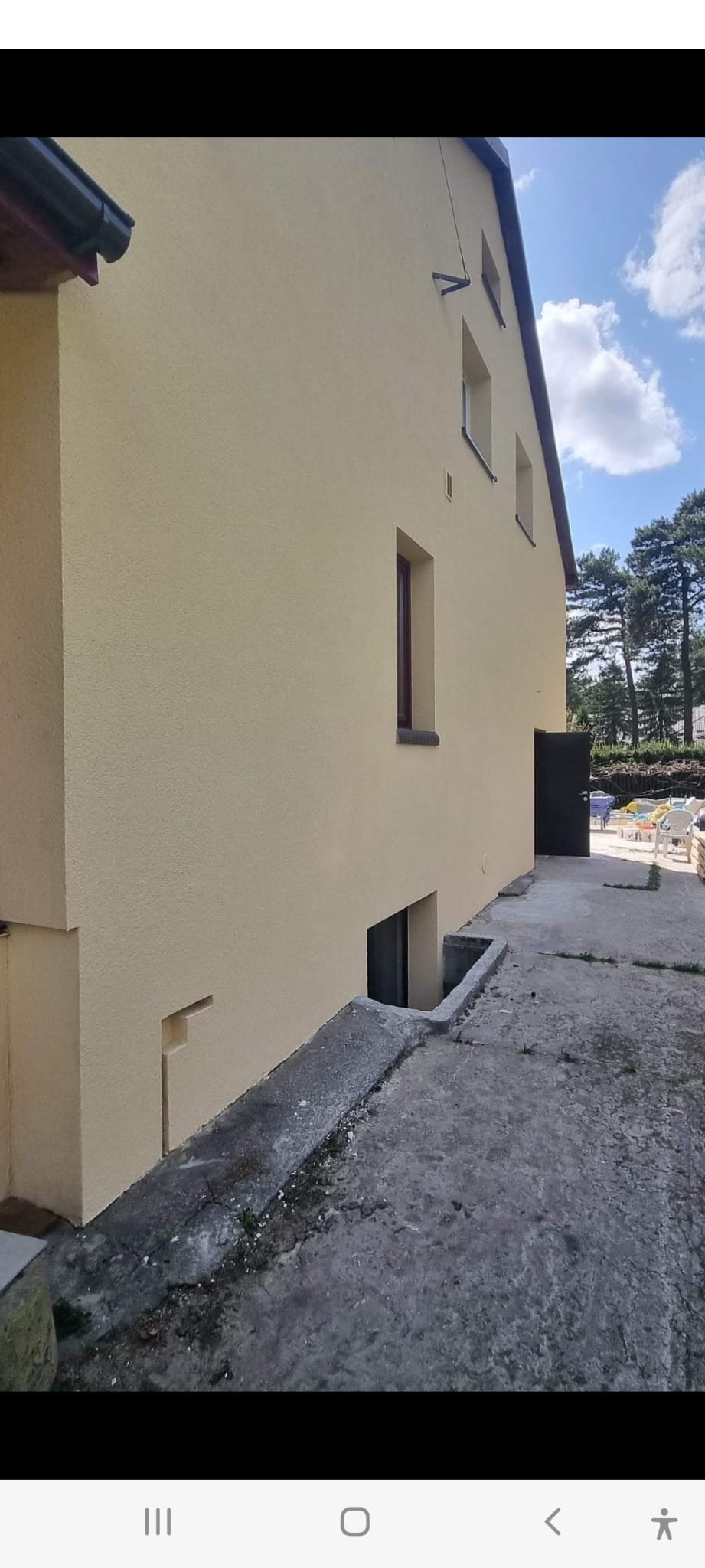 Elewacja domu jednorodzinnego po nałożeniu jasnego tynku, widoczne okna, drzwi wejściowe do piwnicy i fragment chodnika.