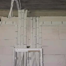 Białe przewody elektryczne ułożone równolegle i przymocowane metalowymi uchwytami do surowej ściany z betonu komórkowego, w tle widoczna puszka rozdzielcza z listwą zaciskową.