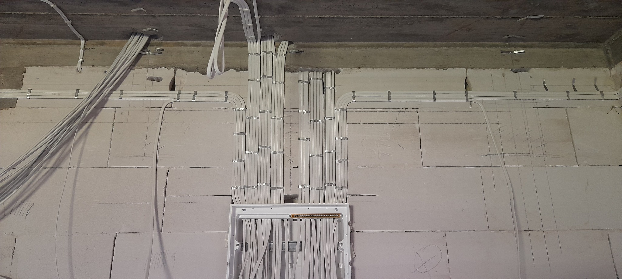 Białe przewody elektryczne ułożone równolegle i przymocowane metalowymi uchwytami do surowej ściany z betonu komórkowego, w tle widoczna puszka rozdzielcza z listwą zaciskową.