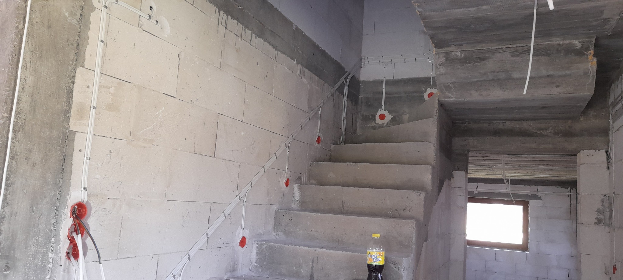 Surowe wnętrze w trakcie prac wykończeniowych: widoczne przewody elektryczne biegnące wzdłuż ściany i schodów, puszki instalacyjne w ścianie, butelka napoju na schodach.