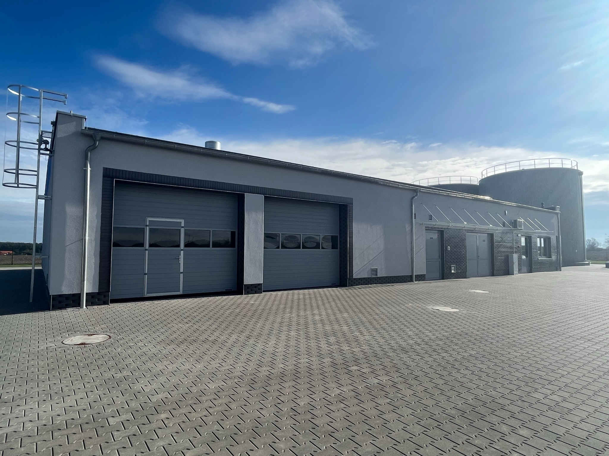 Szara hala przemysłowa z dwoma bramami segmentowymi i stalową drabinką na elewacji, zbiorniki na dachu, wyłożona kostką brukową na zewnątrz, błękitne niebo w tle.