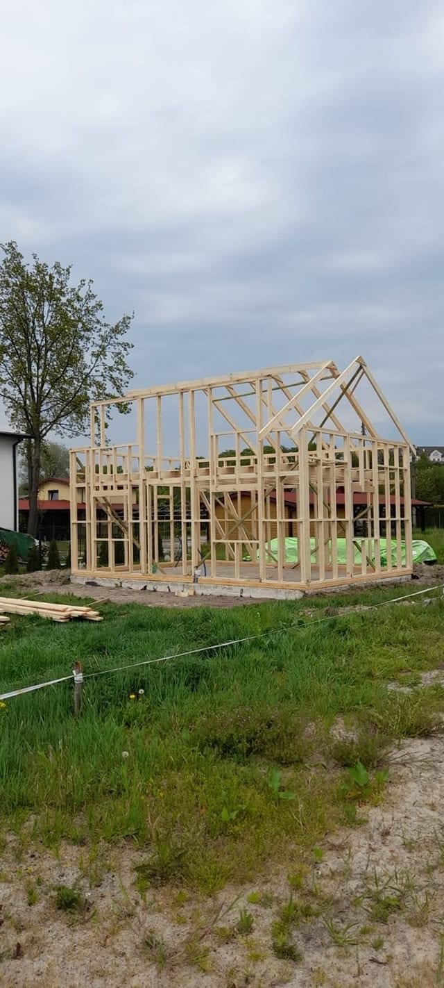 Surowa konstrukcja drewniana domu jednorodzinnego w trakcie budowy, widoczne elementy szkieletu i więźby dachowej na tle pochmurnego nieba i zielonej trawy.