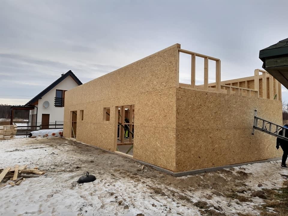 Budowa budynku jednorodzinnego dwulokalowego w stanie surowym otwartym, ściany z płyt OSB, widoczna konstrukcja szkieletowa dachu, zimowa sceneria z resztkami śniegu.