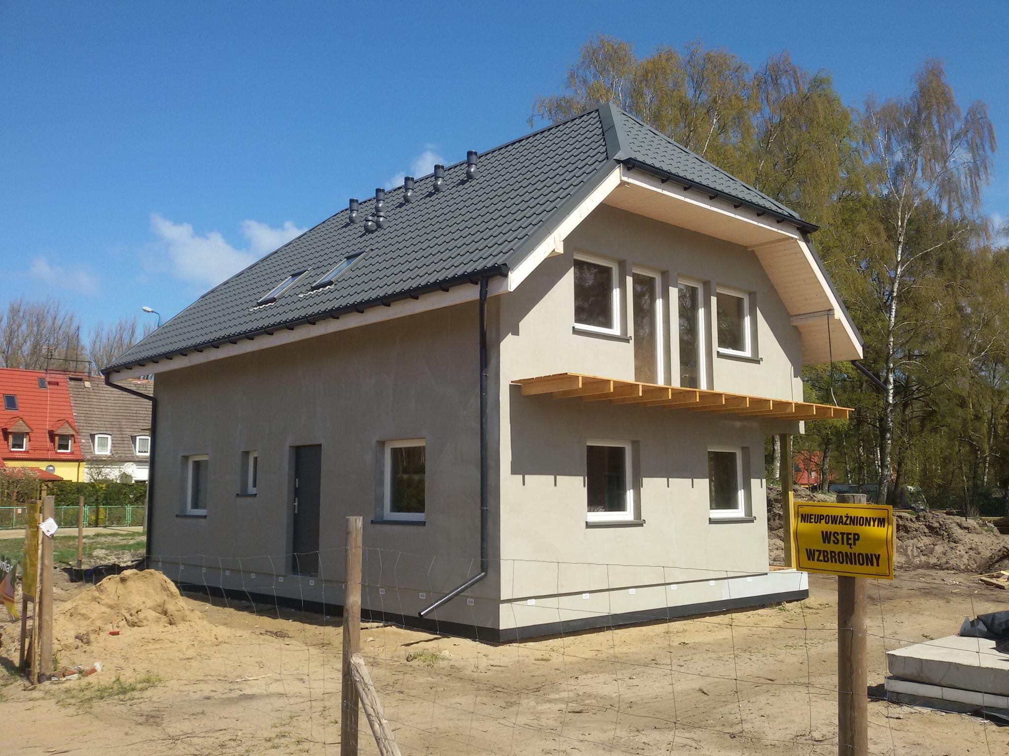 Surowy stan domu jednorodzinnego z szarą blachodachówką, oknami bez parapetów i drewnianą konstrukcją zadaszenia werandy, otoczony piaskiem i ogrodzeniem z siatki.