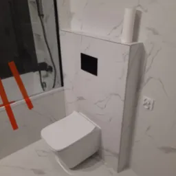 Nowoczesna łazienka z białą armaturą i marmurowymi płytkami, widoczna podtynkowa spłuczka i rolka papieru toaletowego, fragment kabiny prysznicowej z czarną armaturą.