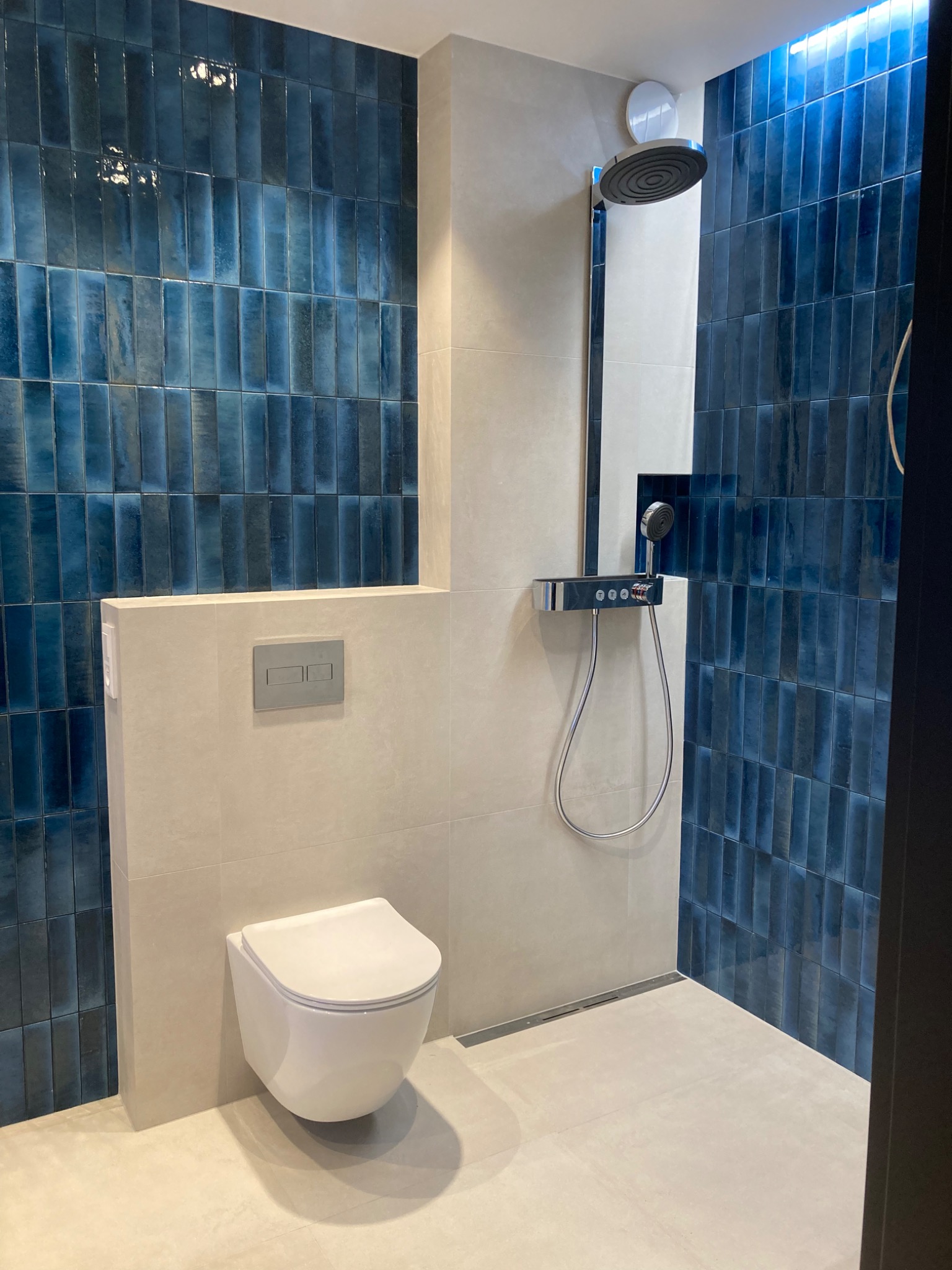 Nowoczesna łazienka z toaletą podwieszaną, armaturą prysznicową z deszczownicą i ścianami wyłożonymi pionowymi, błyszczącymi płytkami w odcieniach niebieskiego.