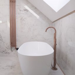 GRESLUX KRZYSZTOF PŁOCINIK - Nowoczesna łazienka na poddaszu z białą wanną wolnostojącą, miedzianą armaturą i grzejnikiem drabinkowym na tle marmurowych płytek i okna dachowego.