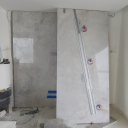 GRESLUX KRZYSZTOF PŁOCINIK - Montaż wielkoformatowych płyt gresowych w łazience. Widoczna szyna tnąca Rubi Slim System i przyssawki. Na podłodze płytki i poziomice. W tle biała ściana i grzejnik.