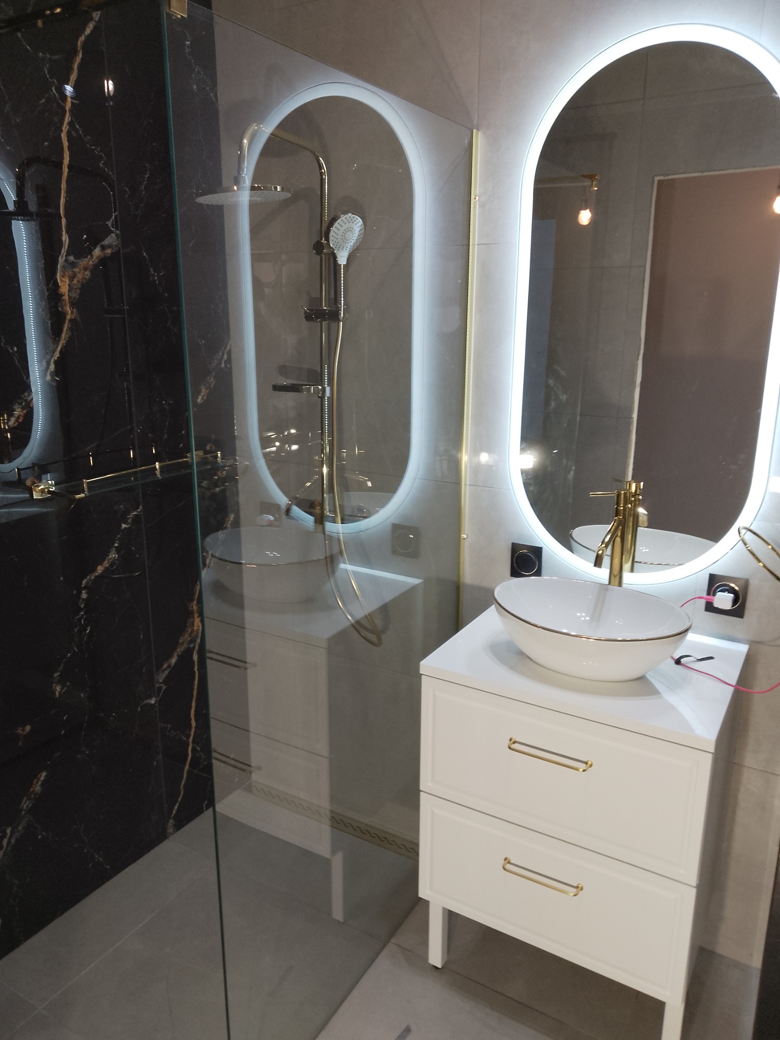 Nowoczesna łazienka z czarną marmurową ścianą, kabiną prysznicową typu walk-in i umywalką nablatową z białym blatem szafki ozdobioną złotymi uchwytami, oświetlone lustra LED w kształcie owalu.