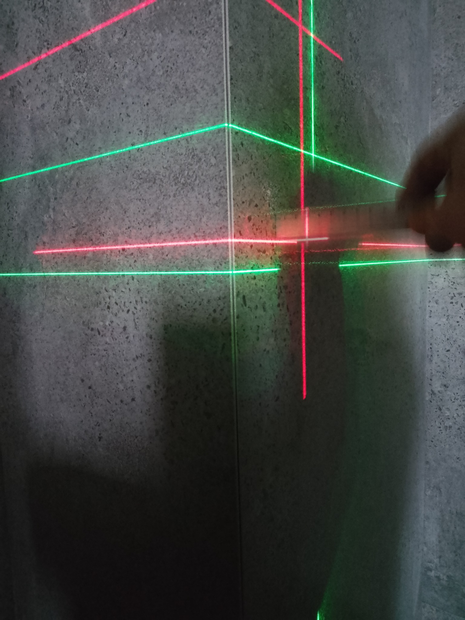 Szara ściana z nałożonymi promieniami lasera krzyżowego w kolorze czerwonym i zielonym, używanego do wyznaczania poziomu, z widoczną dłonią przytrzymującą liniał.
