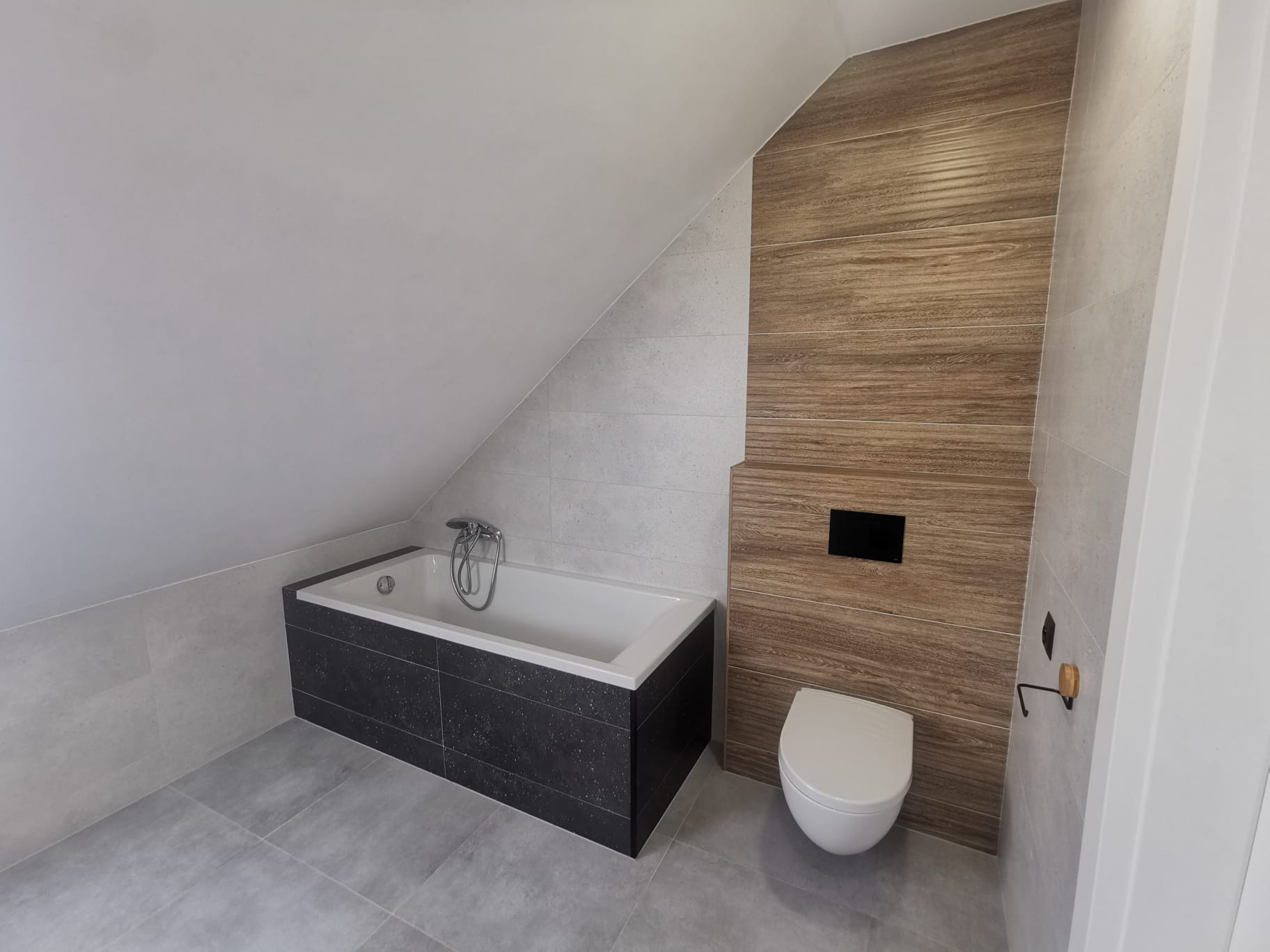Nowoczesna łazienka na poddaszu z wanną obudowaną czarnymi płytkami z brokatem, toaletą podwieszaną i ścianą wyłożoną płytkami imitującymi drewno.