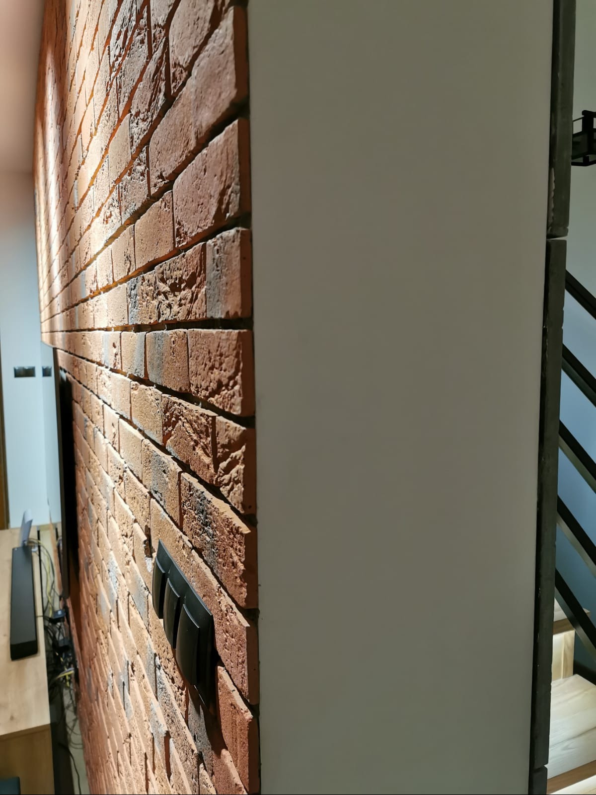 Ściana z czerwonej cegły z czarnymi włącznikami światła, przylegająca do białej, gładkiej ściany w nowoczesnym wnętrzu.