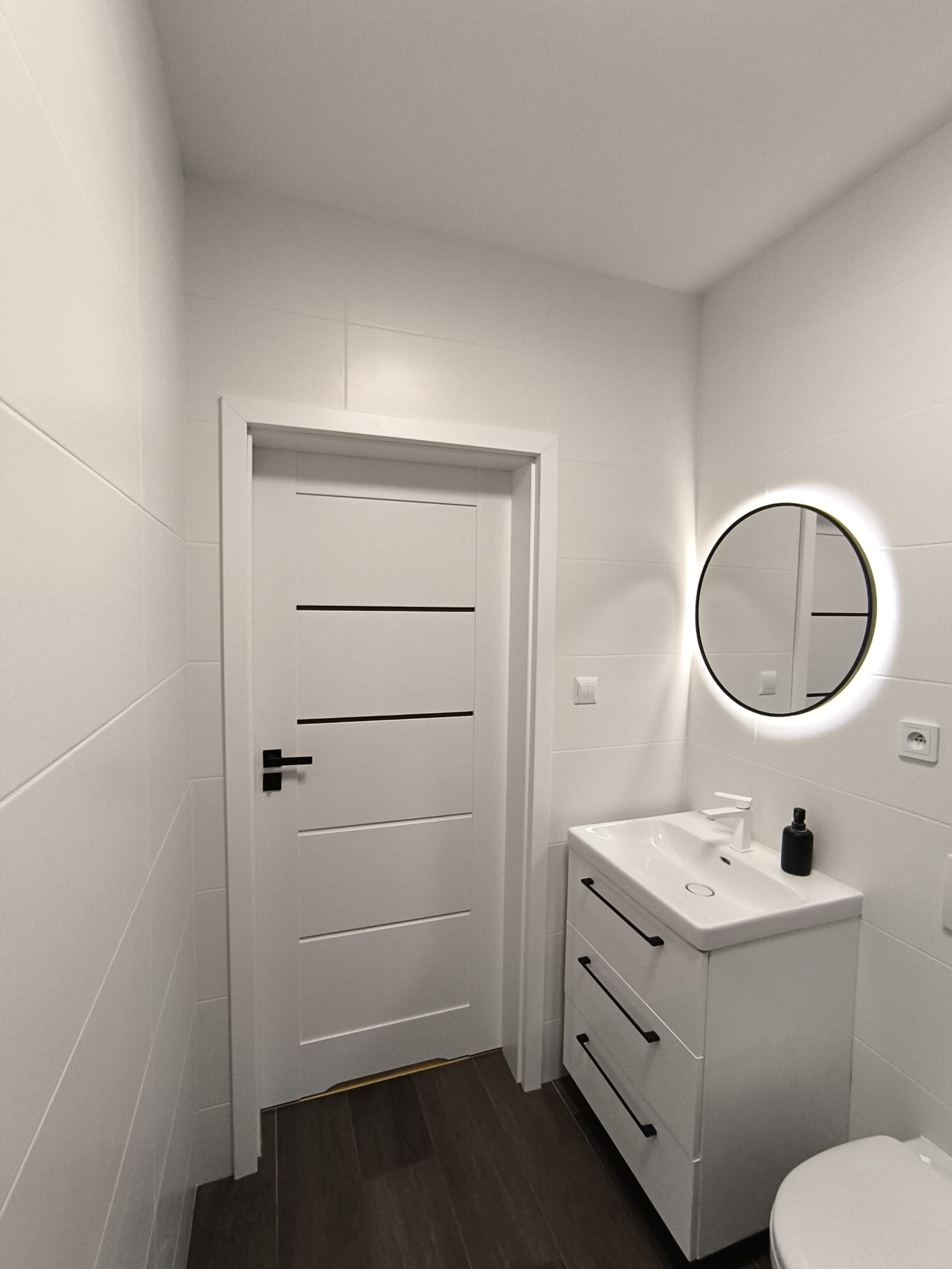 Nowoczesna, minimalistyczna łazienka z białymi płytkami, szafką pod umywalkę z czarnymi uchwytami, okrągłym lustrem z podświetleniem LED i drewnopodobną podłogą.