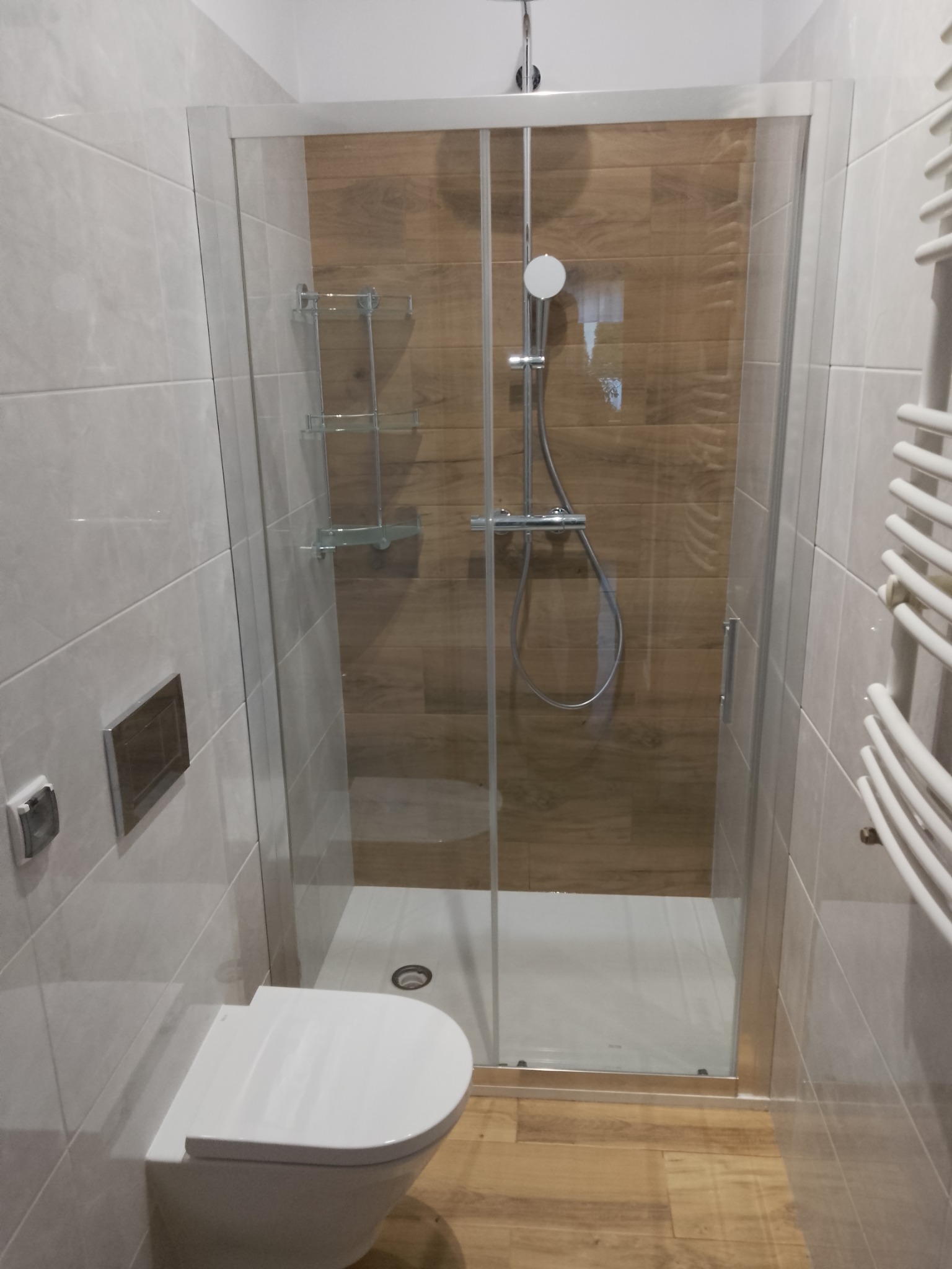 Nowoczesna kabina prysznicowa z drewnopodobnymi płytkami wewnątrz, biała ceramika i grzejnik drabinkowy w łazience wykończonej jasnymi płytkami.