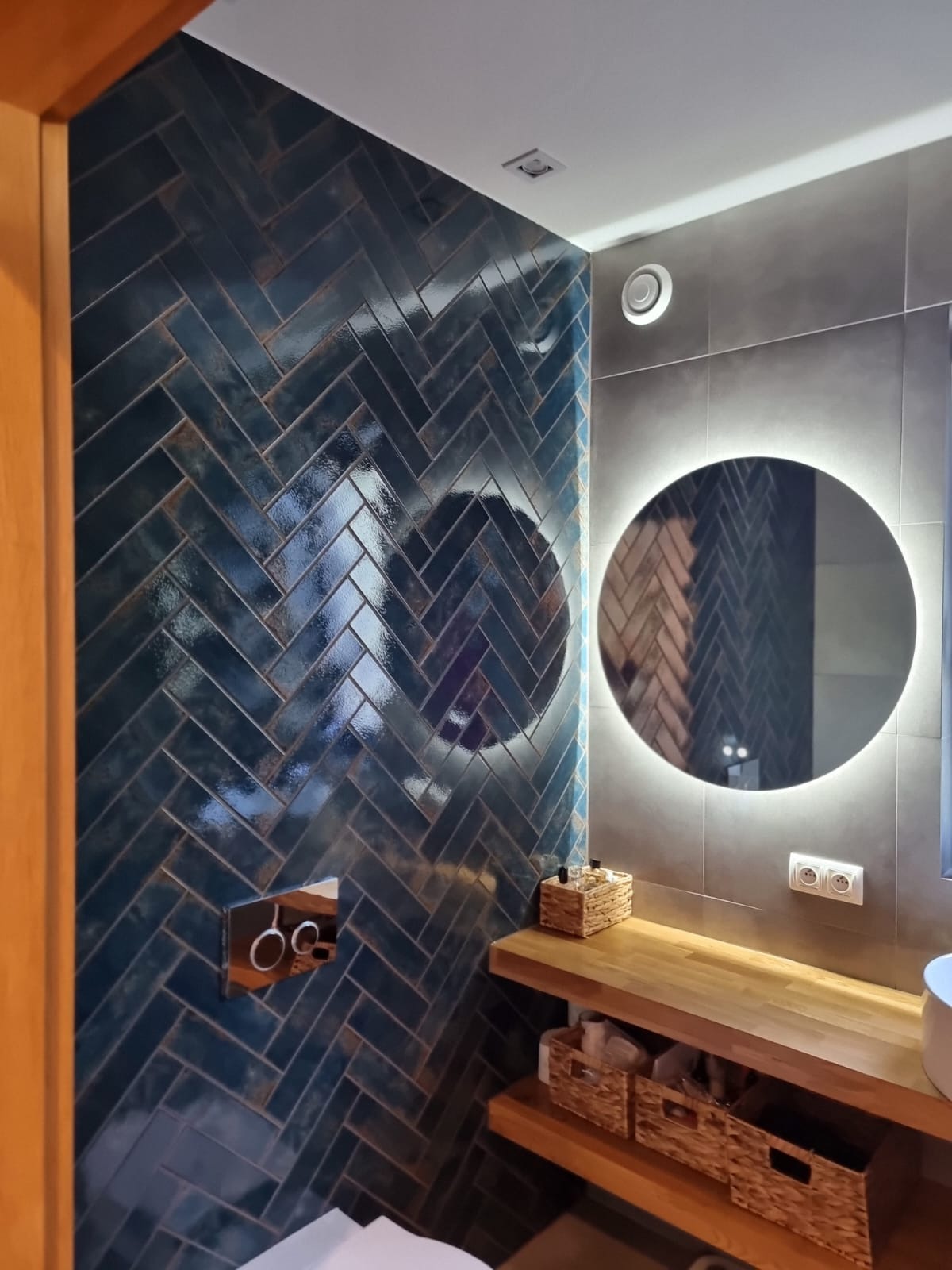 Łazienka z ciemnymi, błyszczącymi płytkami ułożonymi w jodełkę, okrągłe lustro z podświetleniem LED i drewniana półka z koszykami, nowoczesny styl.