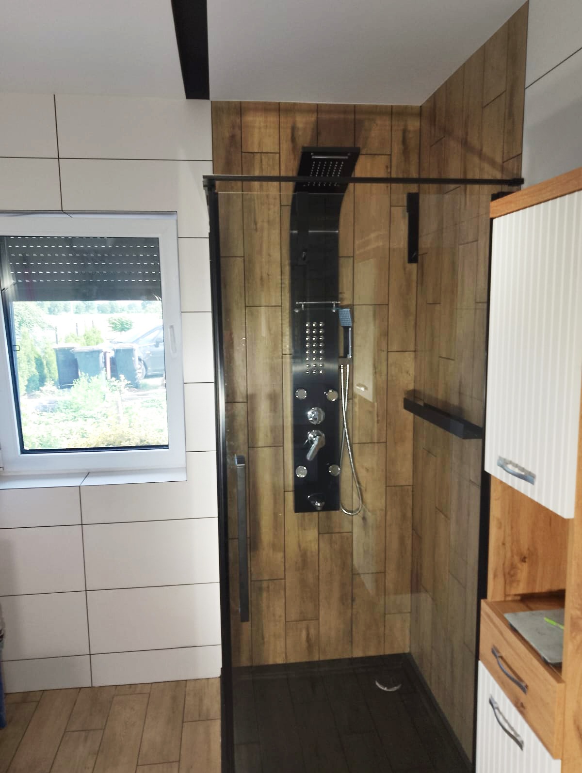 Nowoczesna kabina prysznicowa z panelem natryskowym w łazience wykończonej płytkami imitującymi drewno i białymi kafelkami.