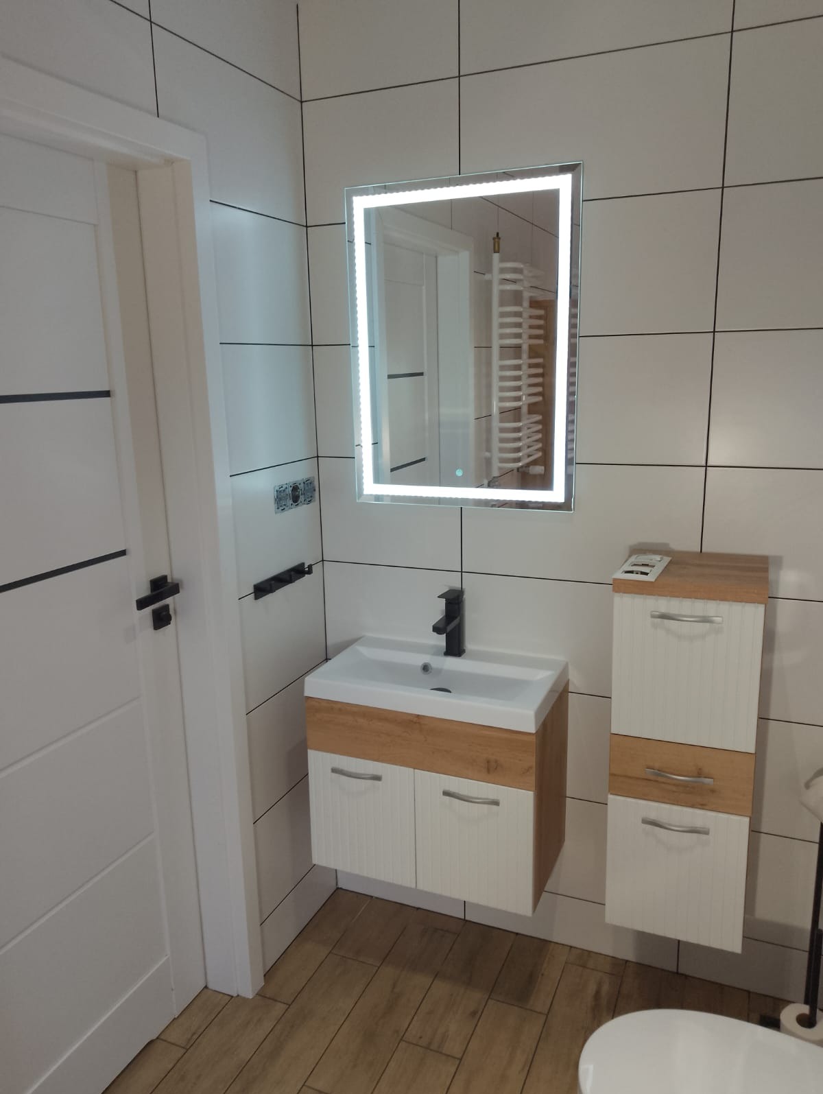 Biała łazienka z drewnianymi akcentami: szafka pod umywalką, słupek i podłoga imitująca drewno. Nad umywalką podświetlane lustro, czarna bateria.