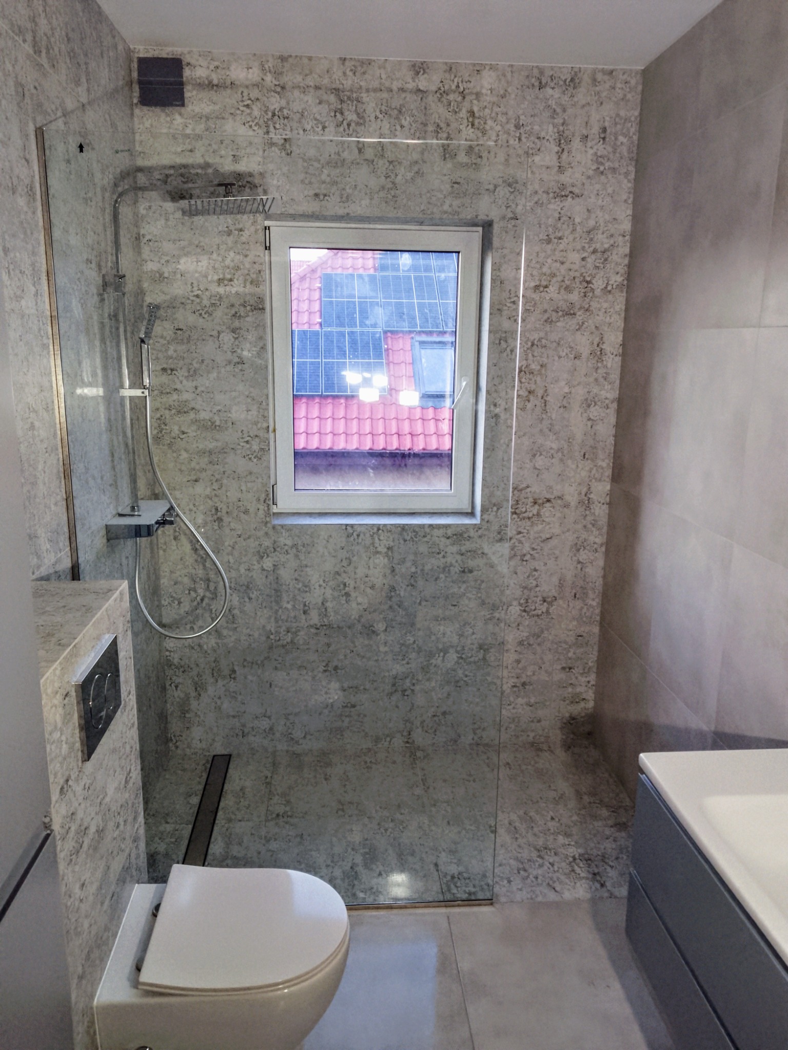 Nowoczesna łazienka z kabiną prysznicową typu walk-in i oknem. Płytki imitujące beton, armatura w kolorze chromu, wisząca toaleta. Widok na dach z panelami słonecznymi.
