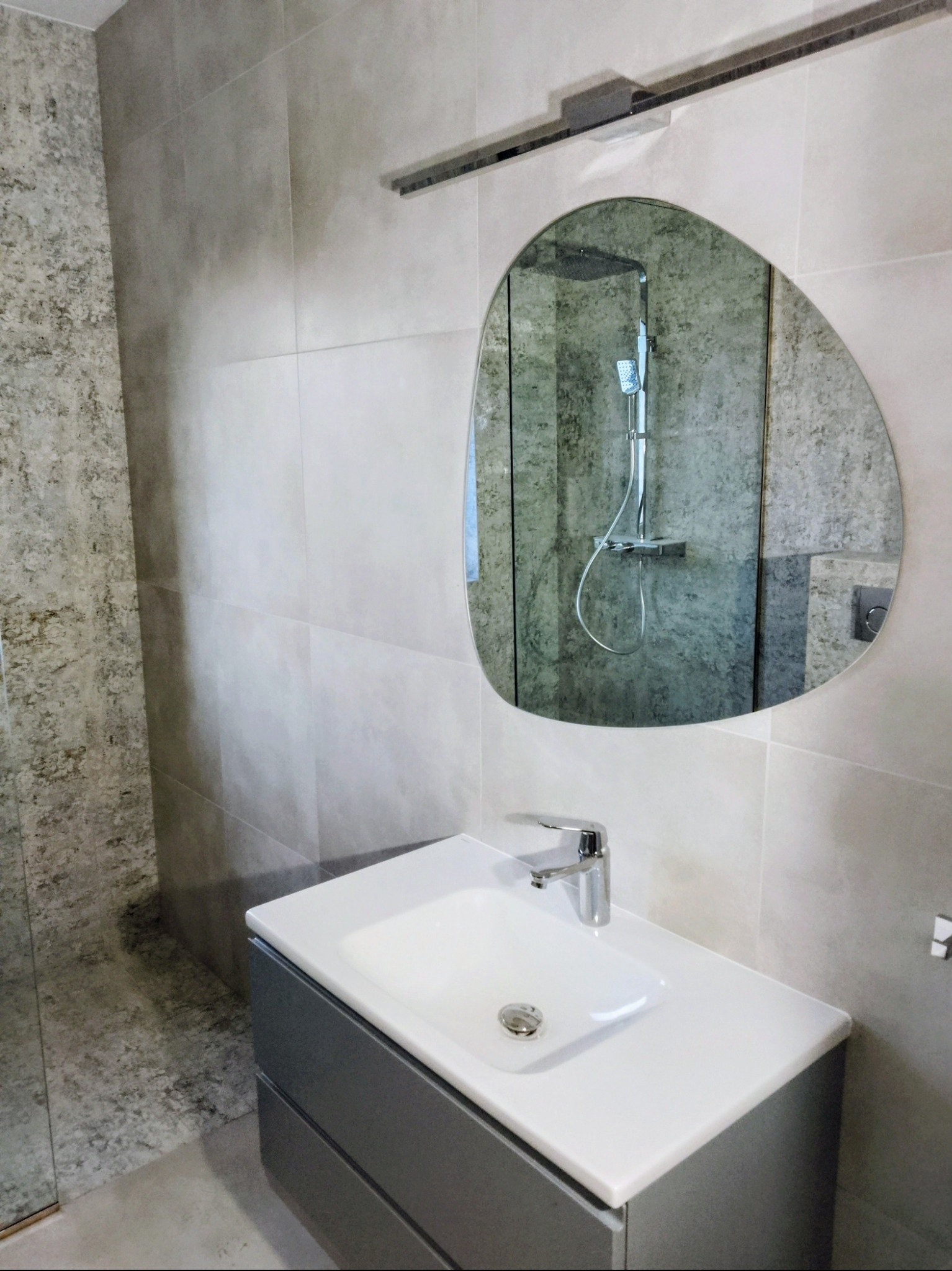 Nowoczesna łazienka w szarościach: asymetryczne lustro, umywalka z szafką, płytki imitujące kamień. Widoczne odbicie kabiny prysznicowej.