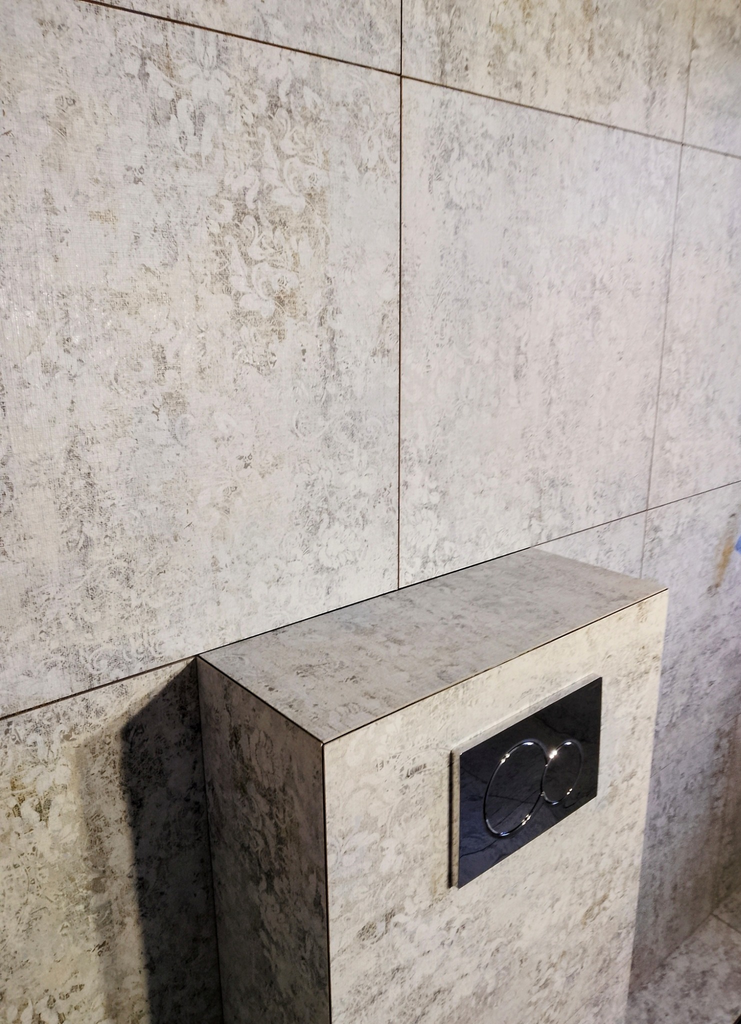 Ściana łazienki wyłożona jasnymi płytkami z delikatnym wzorem, zabudowa stelaża WC z czarnym przyciskiem spłukującym. Minimalistyczny design i stonowana kolorystyka.