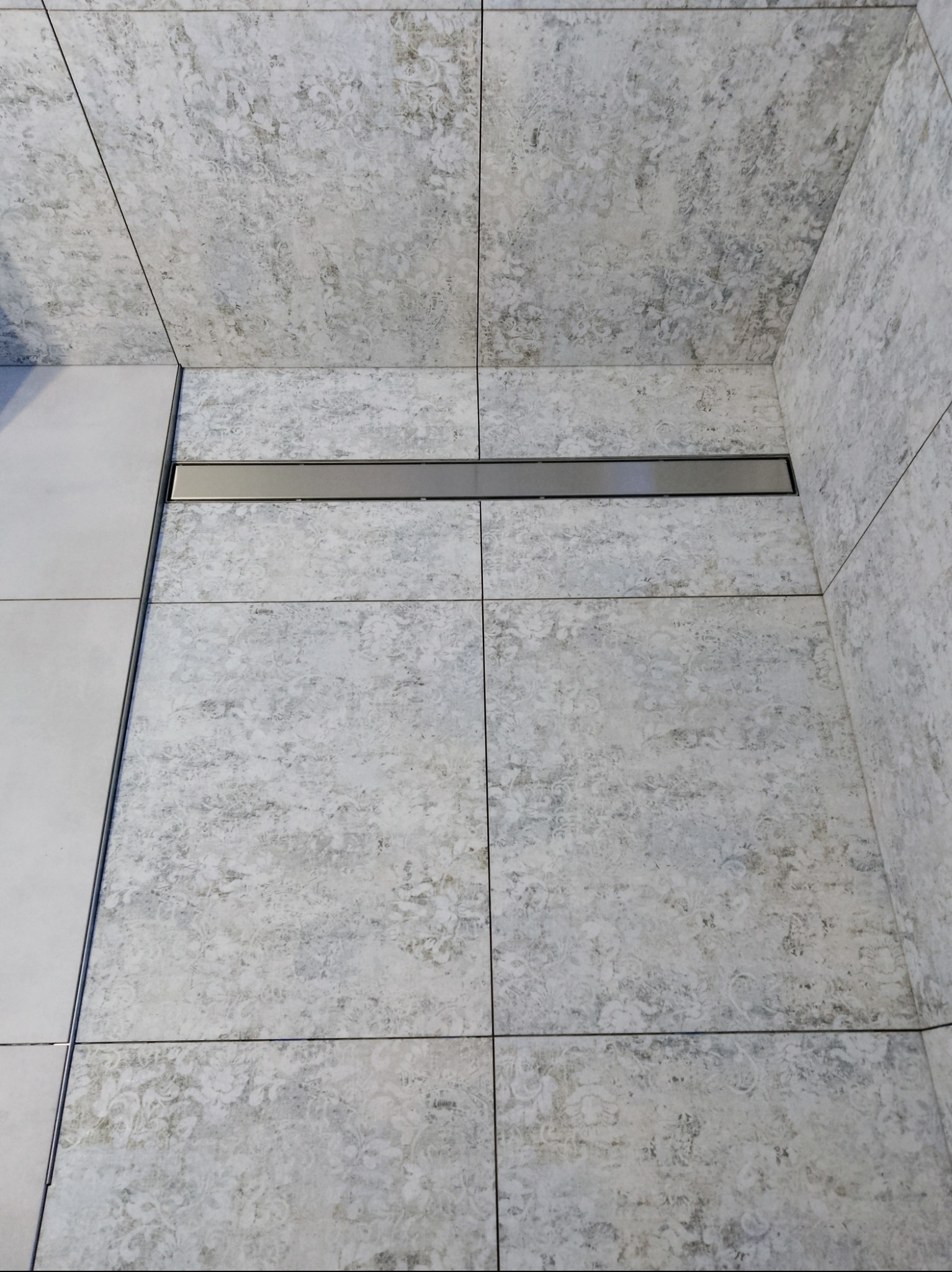 Wykończona łazienka z szarymi płytkami o wzorzystej fakturze i odpływem liniowym. Widok na ścianę i podłogę w narożniku.