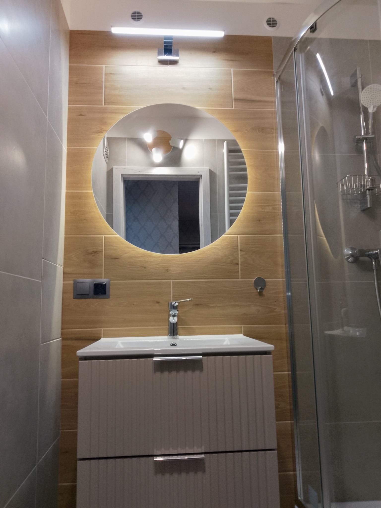 Nowoczesna łazienka: umywalka z szafką, okrągłe lustro z podświetleniem LED na ścianie z płytek imitujących drewno, kabina prysznicowa.