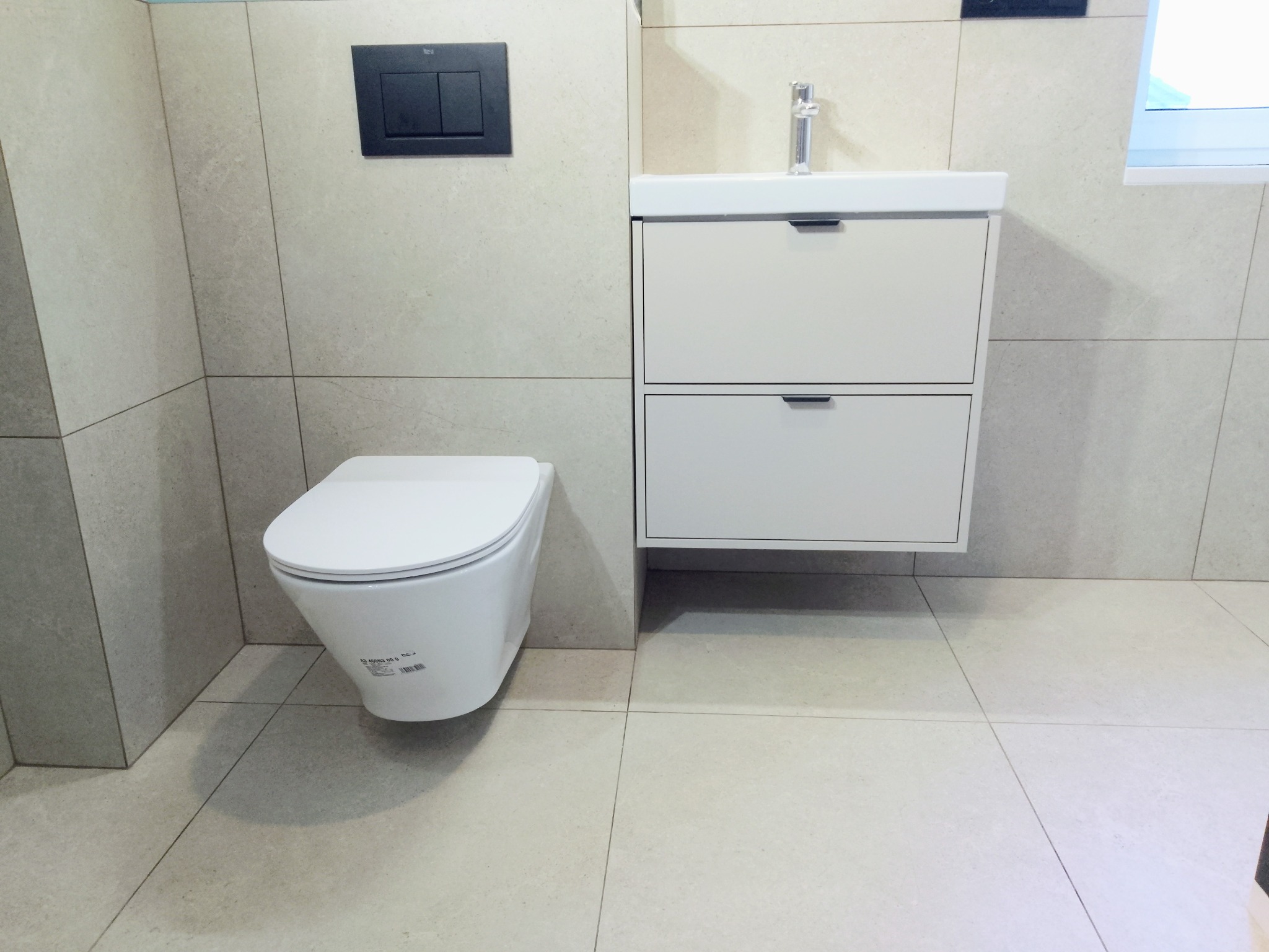 Nowoczesna łazienka z beżowymi płytkami, podwieszaną toaletą i białą szafką z umywalką. Minimalistyczny design, czarne akcenty w postaci przycisku spłukiwania.