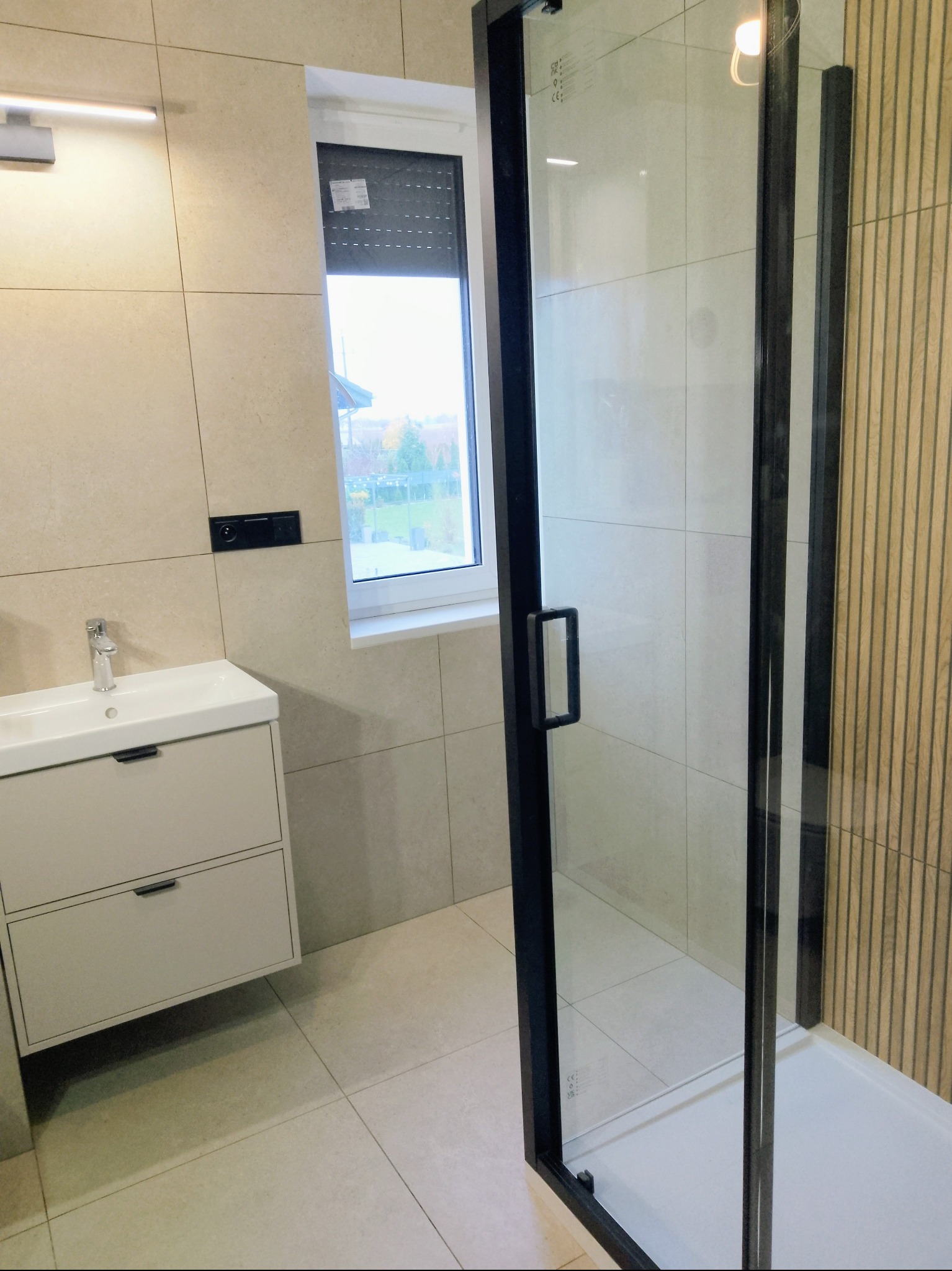 Nowoczesna łazienka z beżowymi płytkami, kabiną prysznicową z czarnymi ramami i szafką pod umywalką. Widok na okno z roletą. Minimalistyczny design.