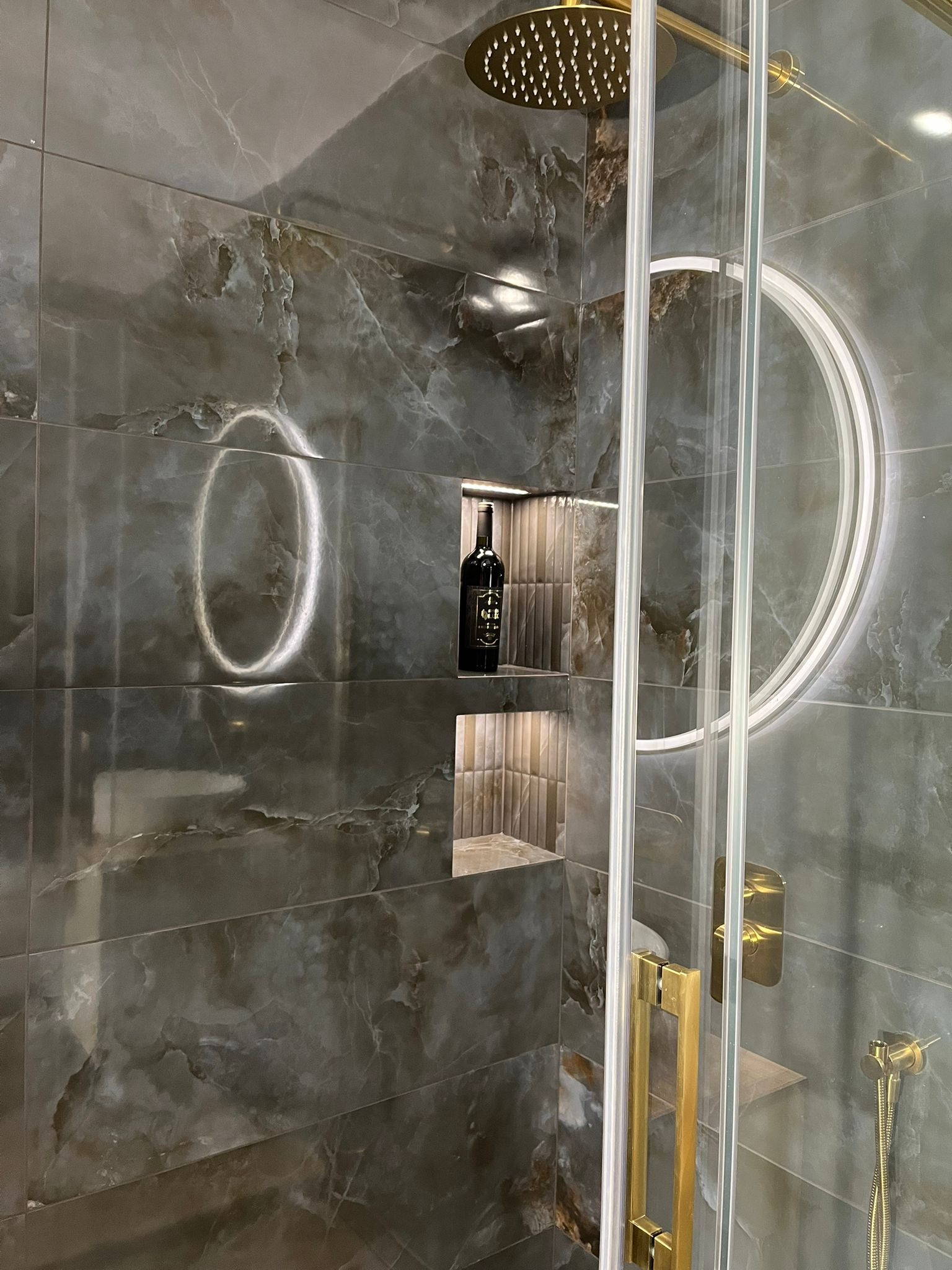 Nowoczesna kabina prysznicowa z ciemnymi płytkami imitującymi marmur, złotą armaturą i podświetlanym lustrem. Wnęka z butelką wina.