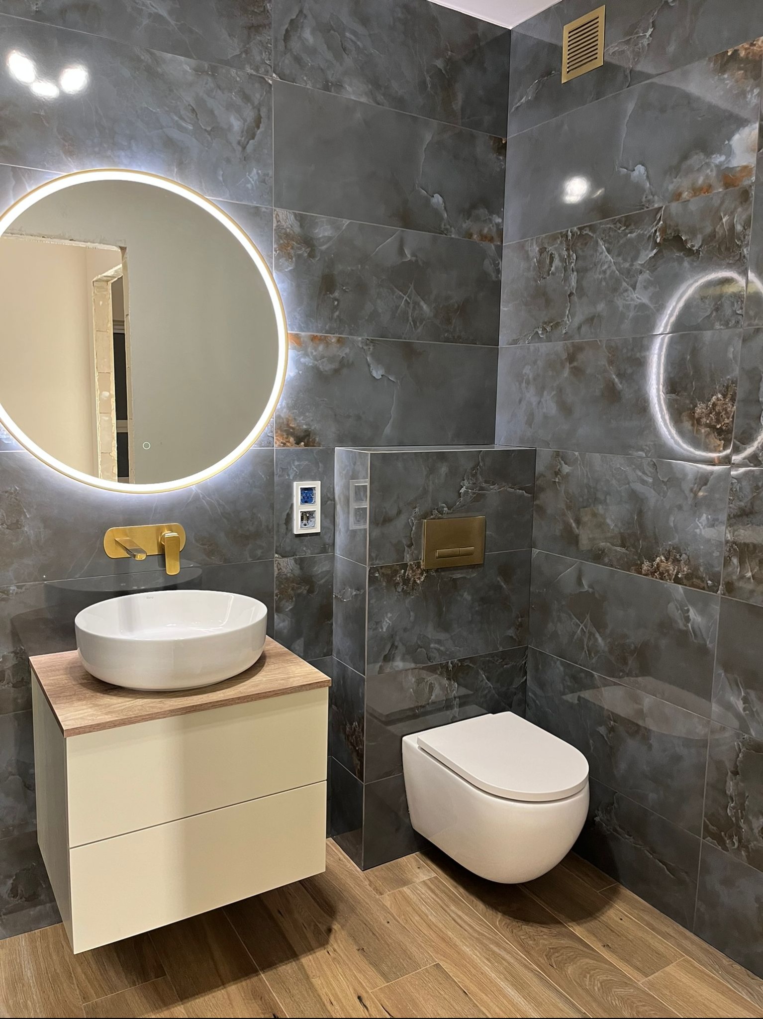 Stylowa łazienka z szarymi płytkami imitującymi marmur, okrągłym lustrem z podświetleniem LED, wiszącą toaletą i drewnopodobną podłogą. Złota armatura dodaje elegancji.