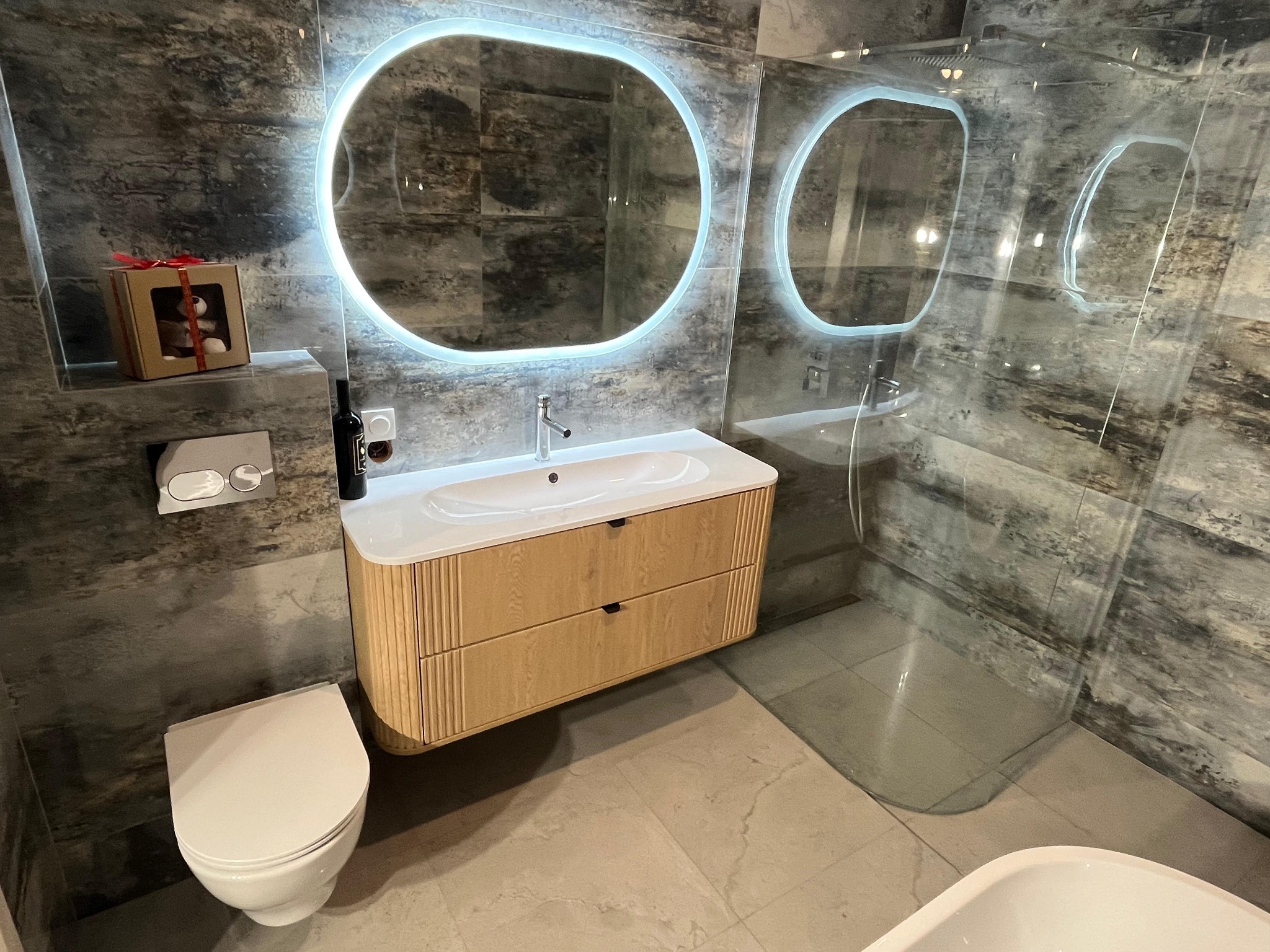 Nowoczesna łazienka z szarymi płytkami imitującymi kamień, podświetlanym lustrem i drewnianą szafką pod umywalką. Na półce prezent i butelka wina.