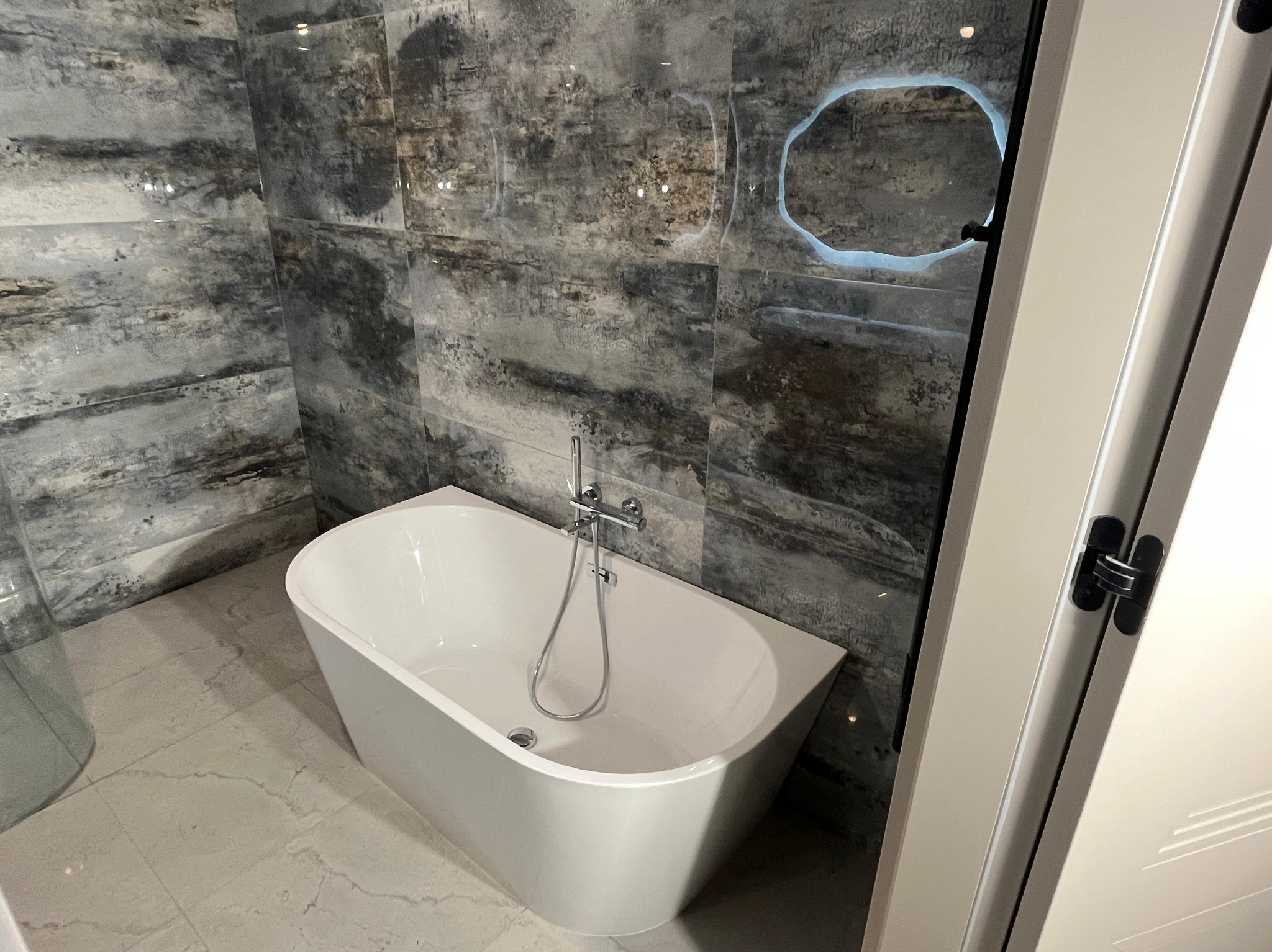 Nowoczesna łazienka z wolnostojącą wanną na tle ściany wyłożonej płytkami imitującymi kamień. Okrągłe lustro z podświetleniem. Minimalistyczna armatura.