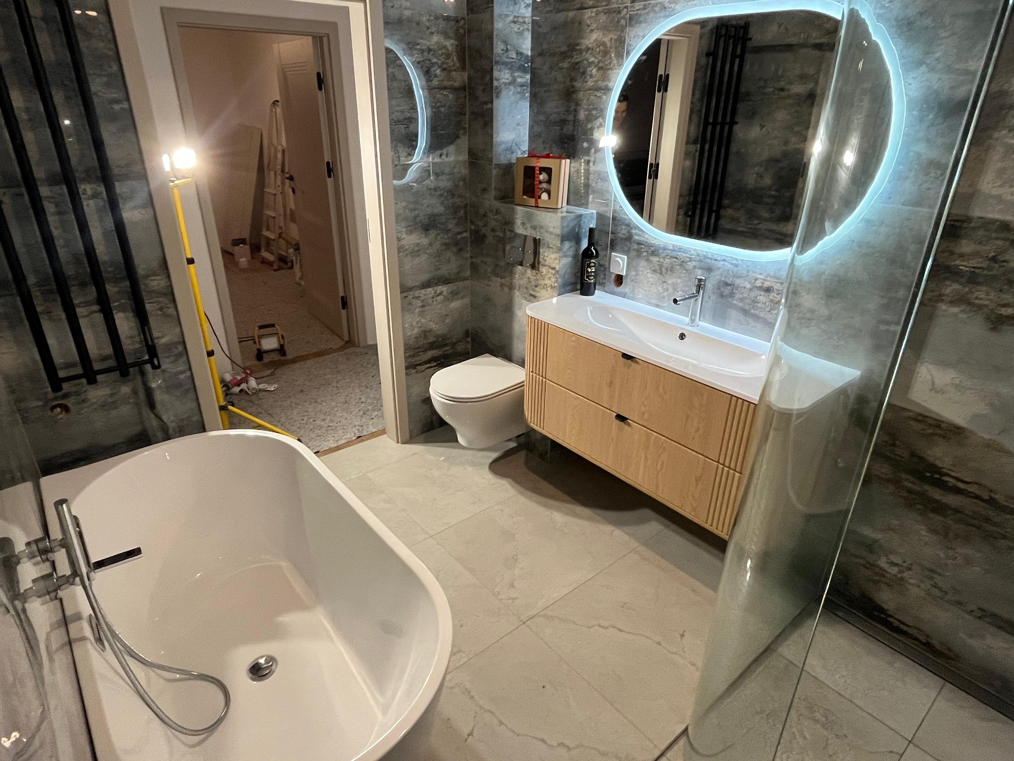 Wykończona łazienka z wanną, umywalką z szafką i podświetlanym lustrem. Widoczna drabina i lampa budowlana sugerują prace wykończeniowe w toku.