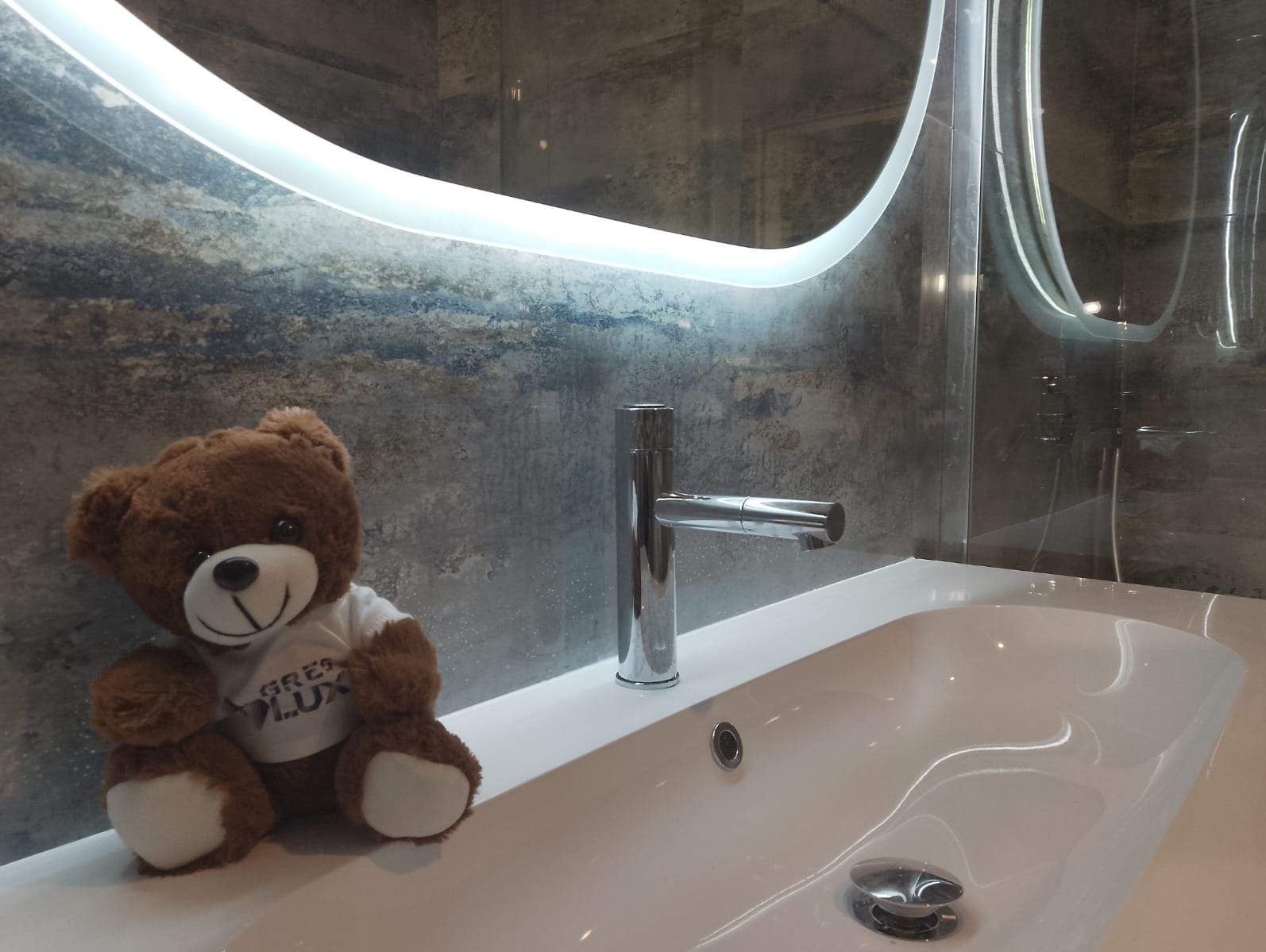 Biała umywalka z chromowaną baterią na tle szarej ściany z efektem betonu i podświetlanego lustra. Pluszowy miś w koszulce z logo firmy dodaje unikalnego charakteru.