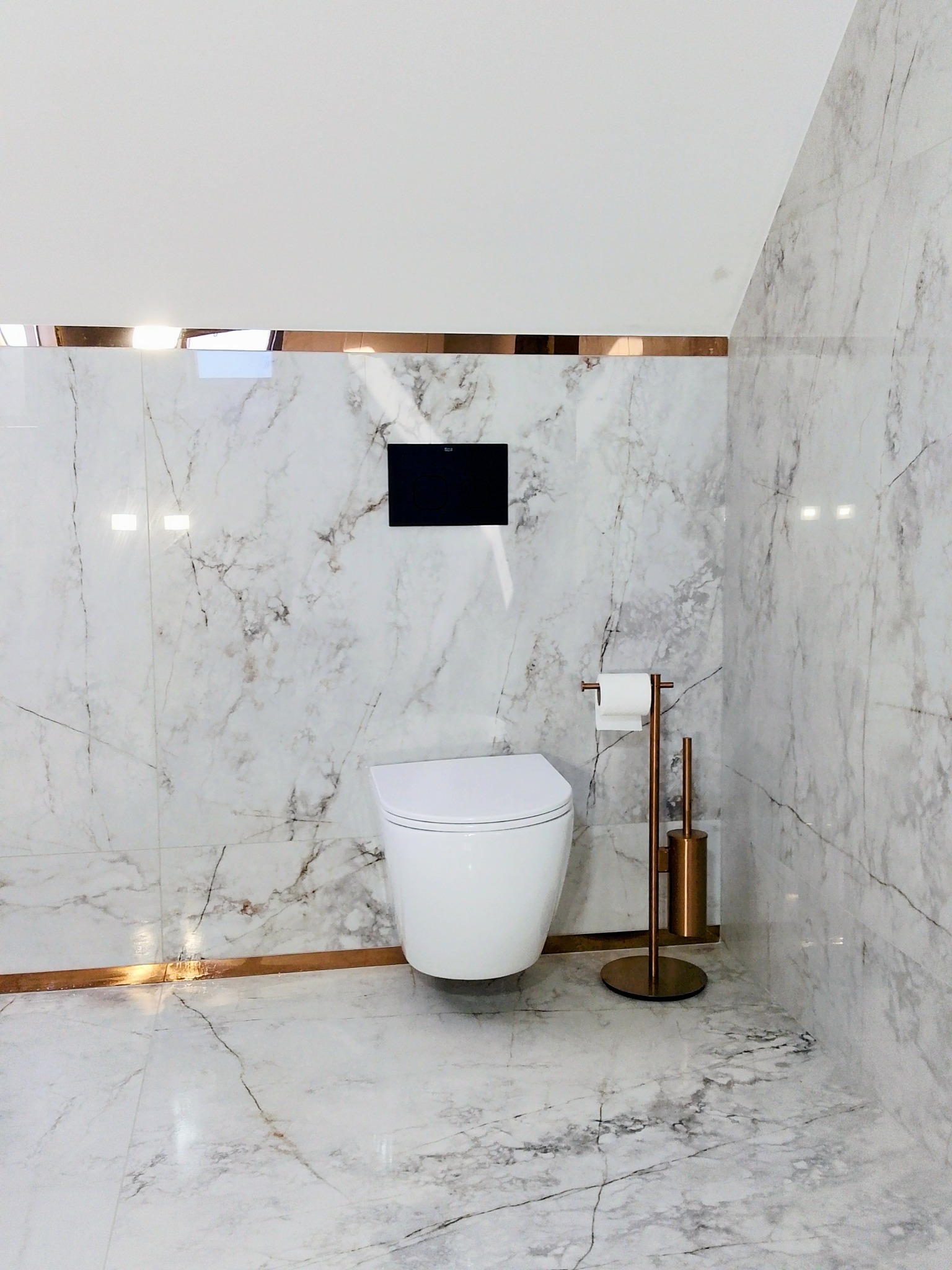 Nowoczesna toaleta podwieszana w łazience wykończonej płytkami imitującymi marmur, z akcentami w kolorze miedzianego złota. Minimalistyczny design.