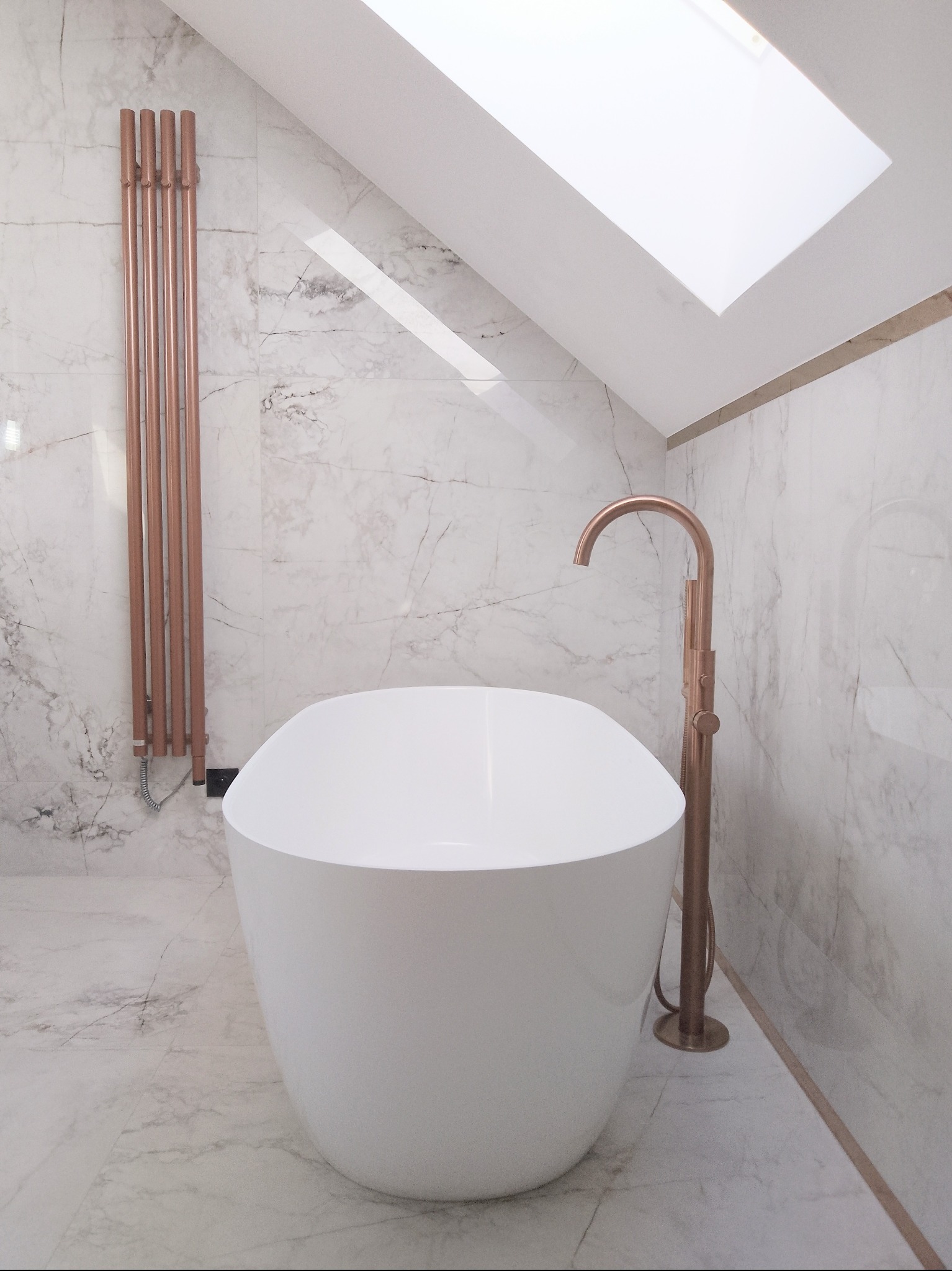 Nowoczesna łazienka na poddaszu z białą wanną wolnostojącą, miedzianą armaturą i grzejnikiem drabinkowym na tle marmurowych płytek i okna dachowego.