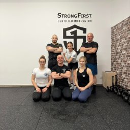 Akademia Sprawności - Grupa sześciu osób w strojach sportowych pozuje w siłowni na tle ściany z logo StrongFirst Certified Instructor, obok widoczne kettlebells.